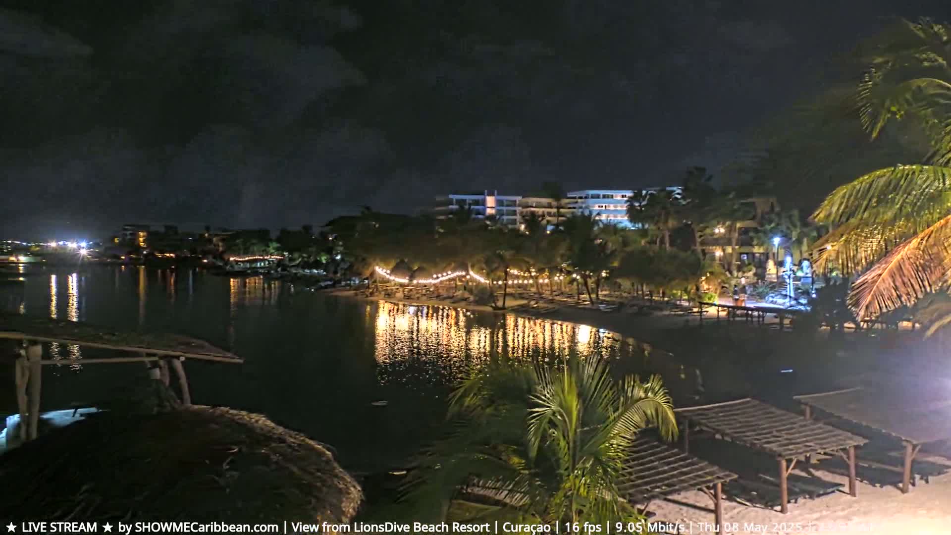 A nighttime view of a calm body of water reflecting the lights of a resort's beach and buildings, with palm trees and outdoor structures visible along the shore.