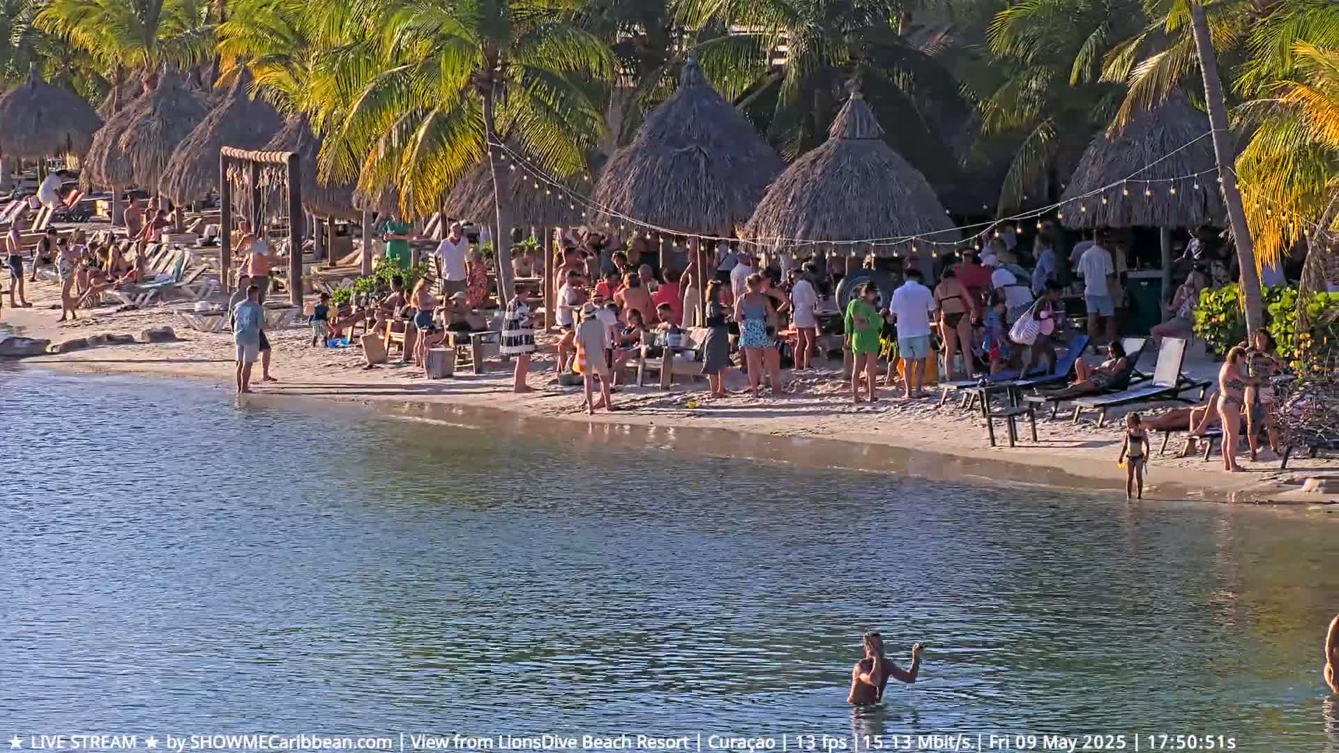 A sandy beach with many people under thatched-roof huts next to calm water.
