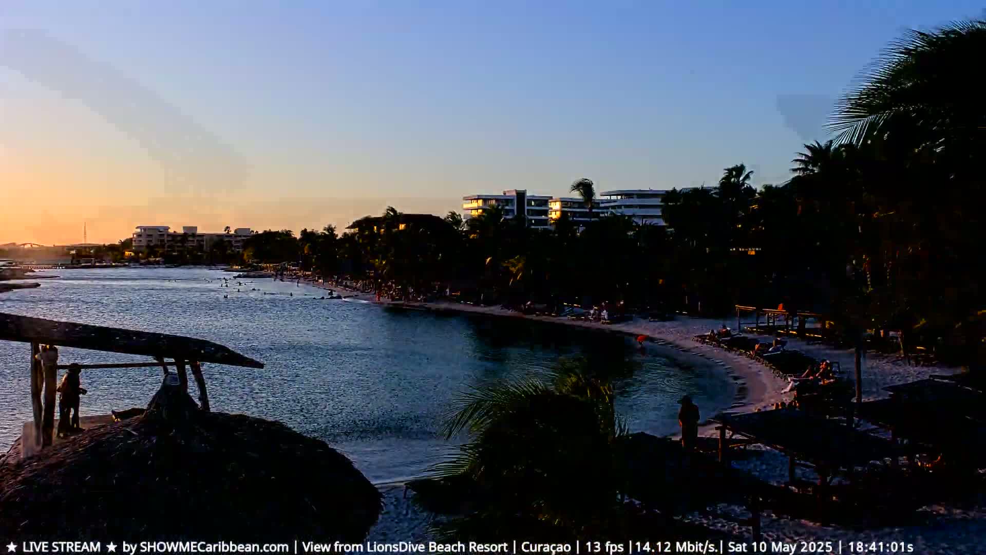 A tranquil sunset scene shows a calm body of water, a sandy beach with people, and resort buildings silhouetted against the twilight sky.