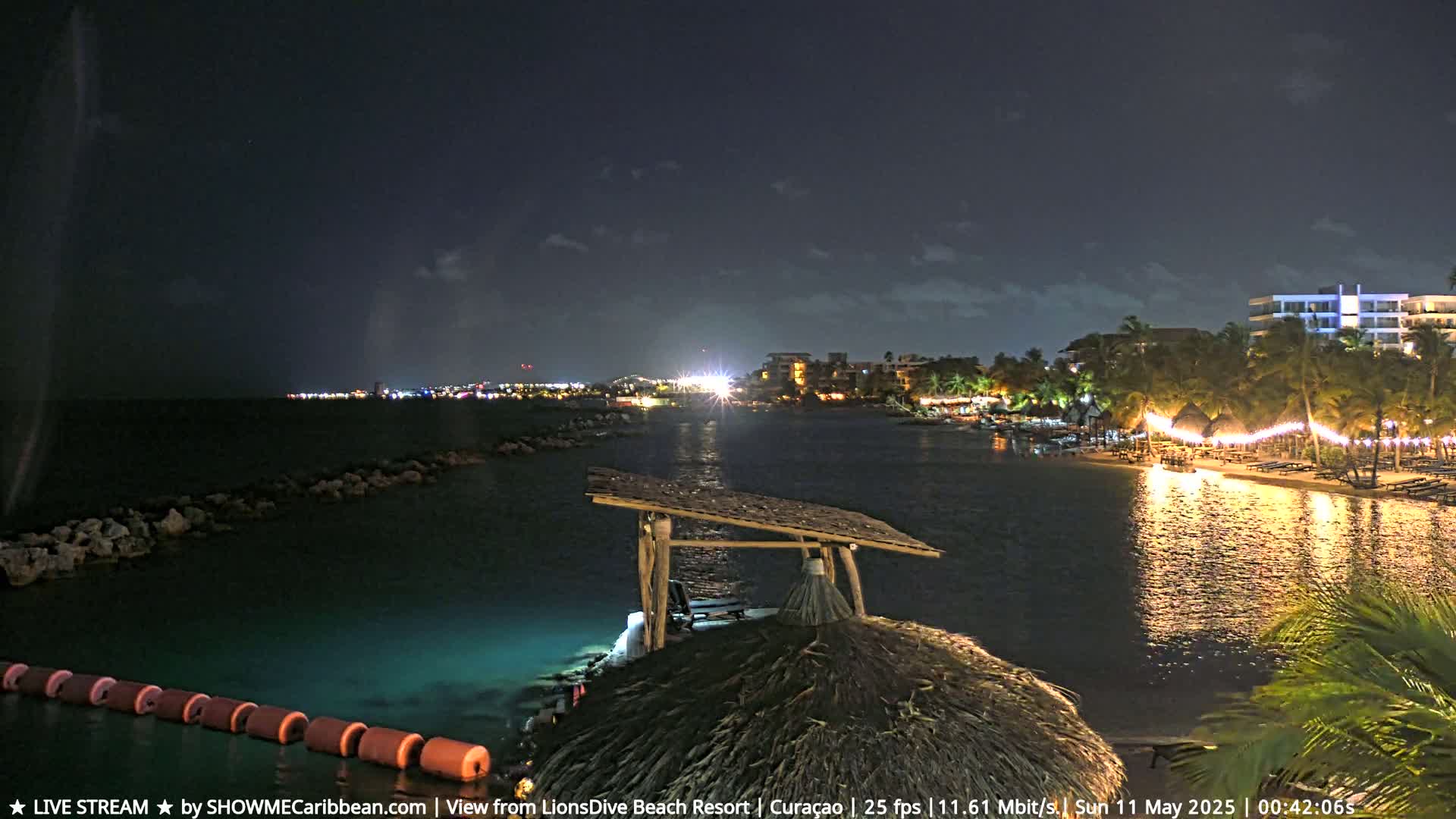 A nighttime view of a calm, illuminated body of water shows a beach with lights strung along it, a resort in the background, and a thatched-roof structure in the foreground.