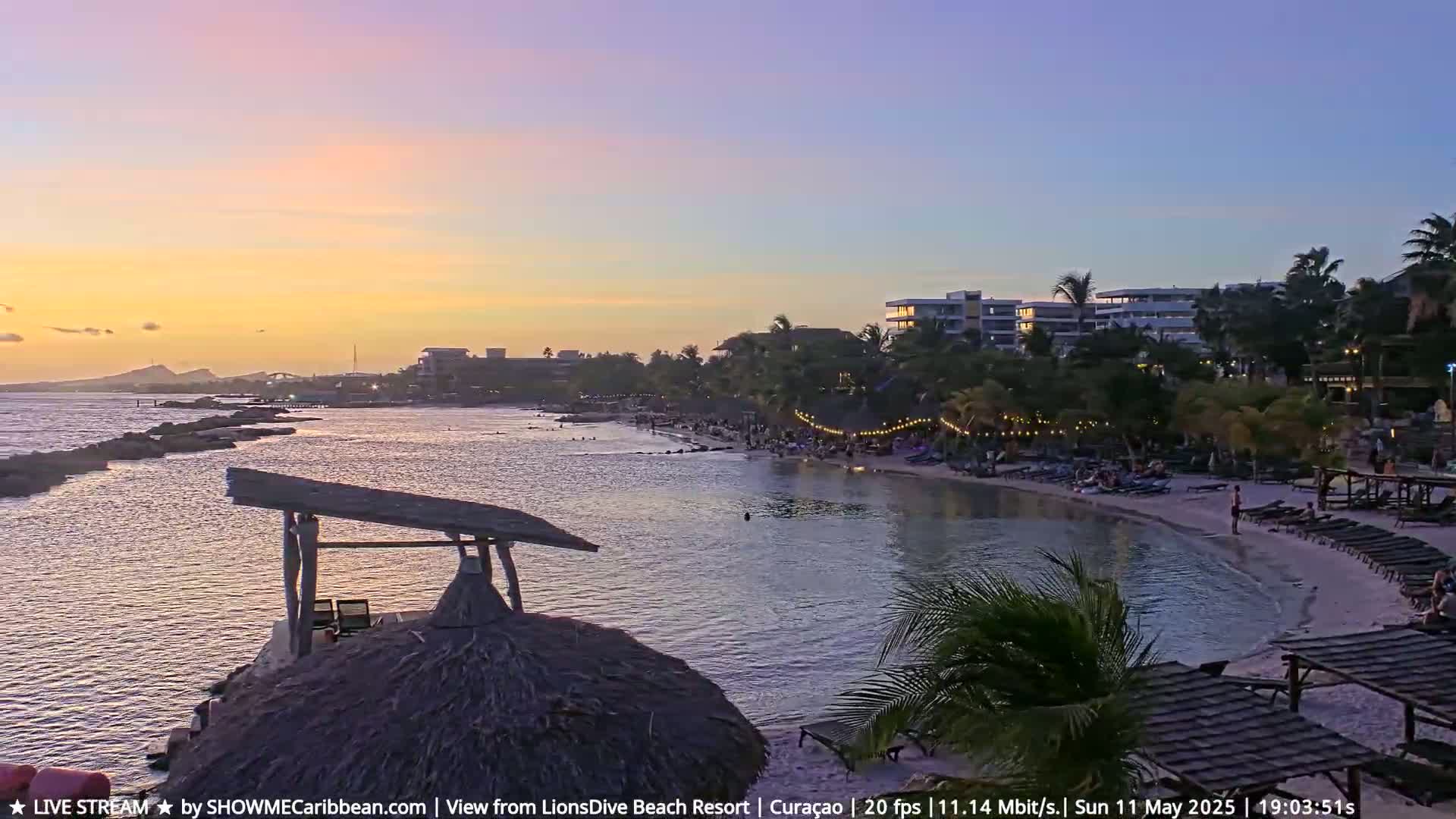 A tranquil sunset over a calm bay, with a sandy beach lined with lounge chairs and palm trees, and buildings visible in the background.