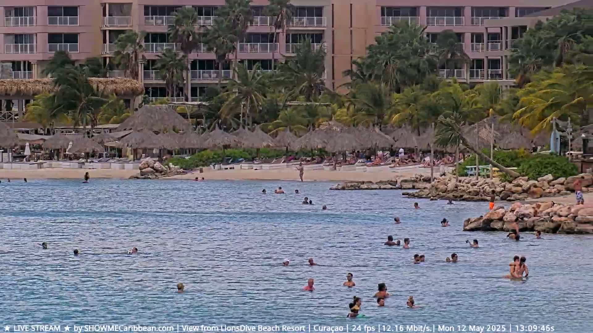 A calm, partly cloudy day shows many people swimming and sunbathing at a beach with thatched-roof umbrellas and a resort building in the background.