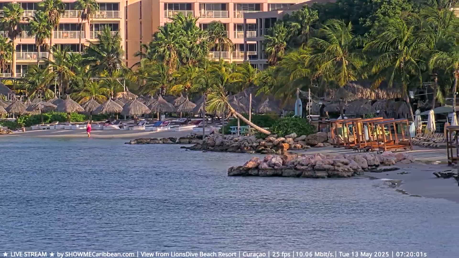A sandy beach with palm trees, thatch-roofed umbrellas, and lounge chairs is situated in front of a multi-story building; a person walks along the shore near calm water.