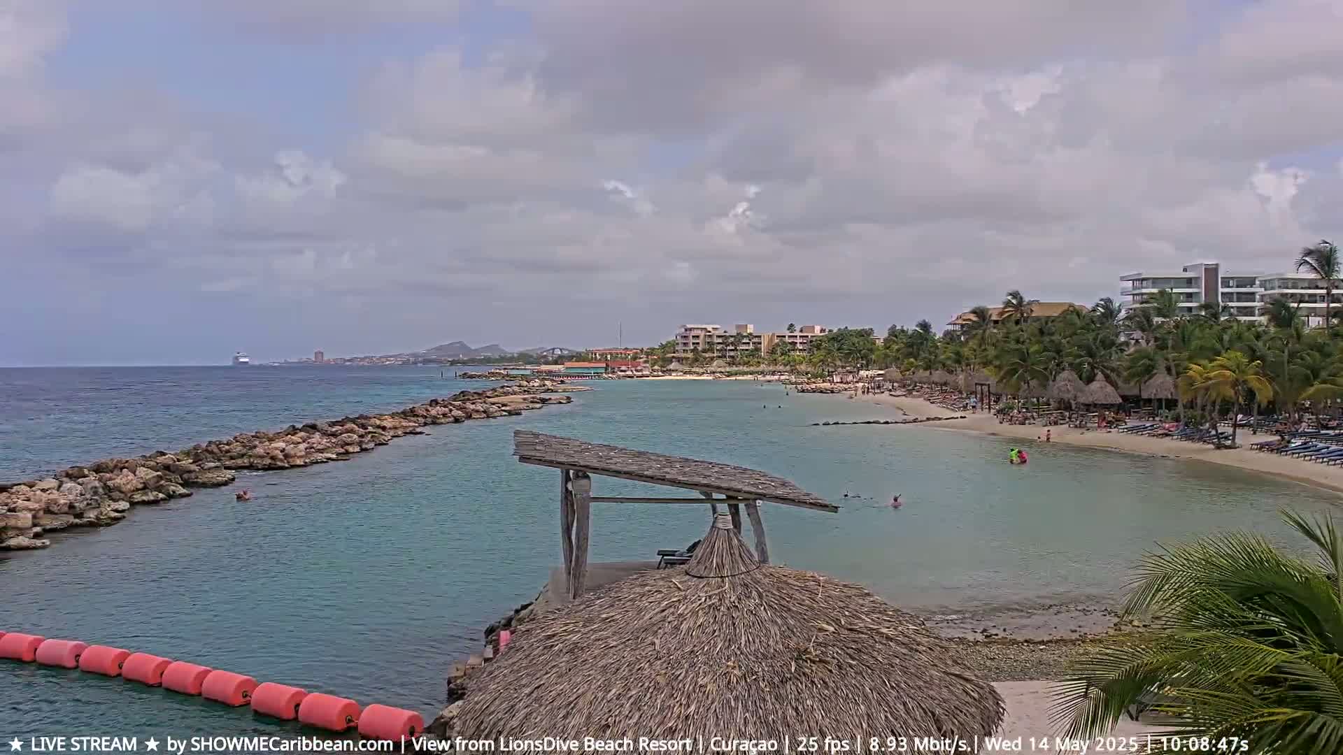 A partly cloudy day at a tropical beach resort shows calm turquoise water, a sandy shore with lounge chairs and people, and a distant cruise ship.