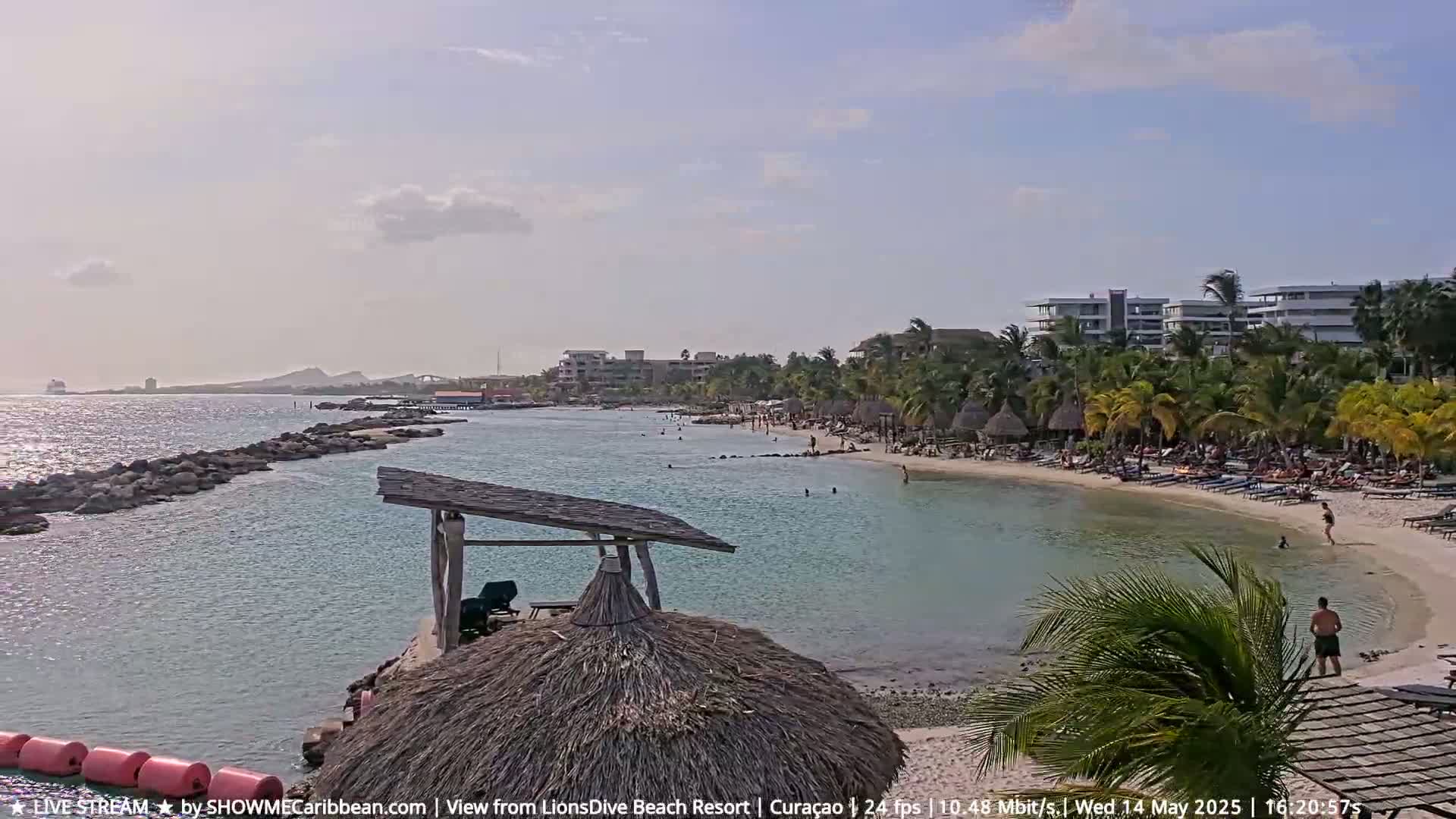 A partly sunny day shows a sandy beach with people sunbathing and swimming in calm turquoise water, backed by palm trees, beach huts, and buildings.