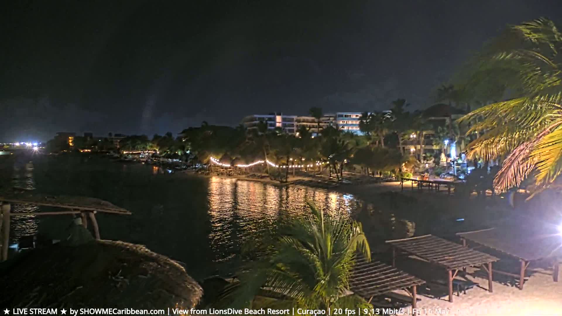 A nighttime view of a calm body of water reflecting lights from a beachside resort and palm trees, under a clear, dark sky.