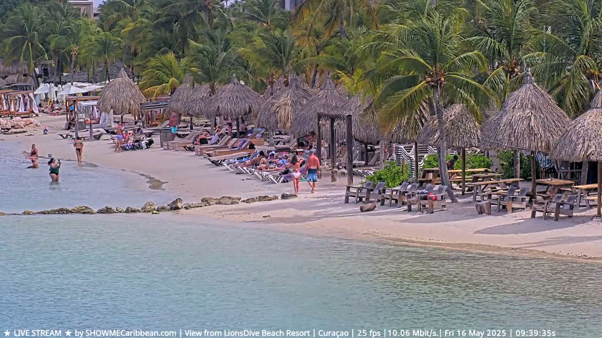 A sandy beach with numerous thatched umbrellas, lounge chairs, and people relaxing and swimming in calm, clear water under a sunny sky, surrounded by lush palm trees.