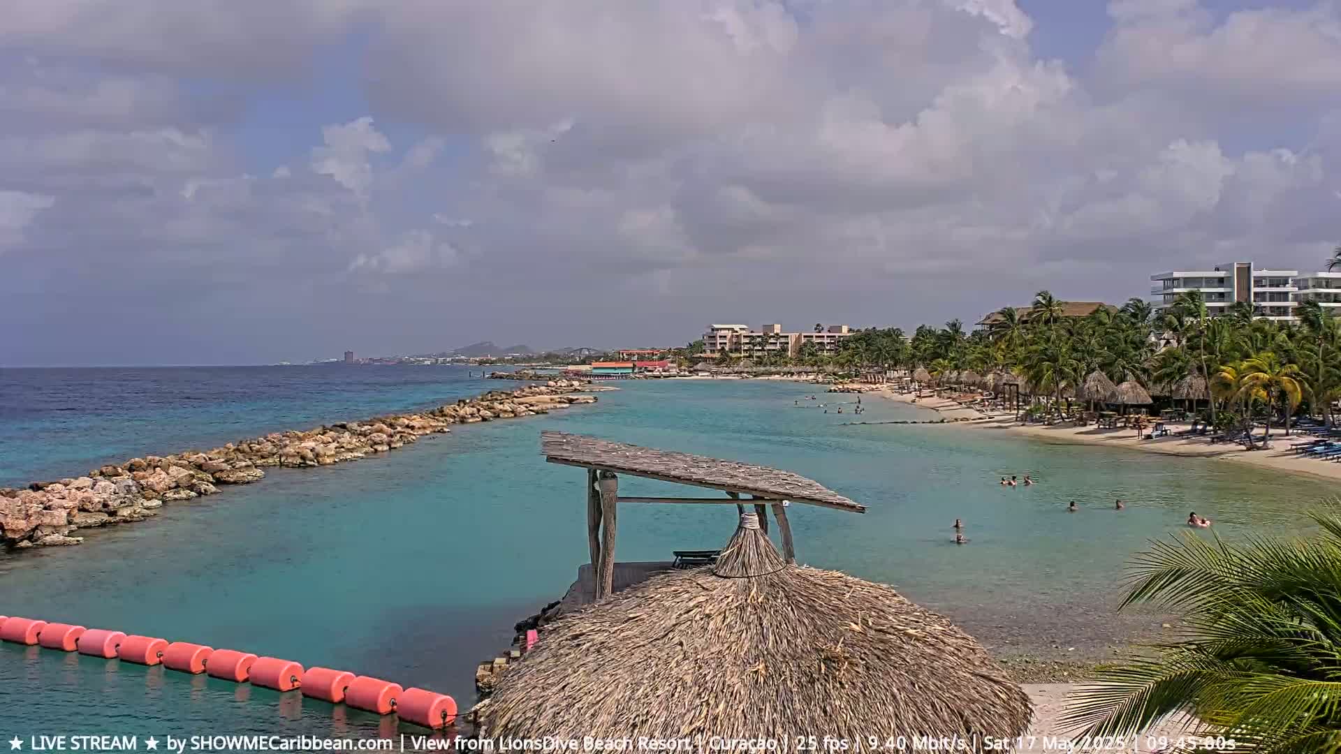 A partly cloudy day at a tropical beach shows calm, clear turquoise water, a sandy shore lined with palm trees and beach umbrellas, and a rock jetty extending into the ocean.