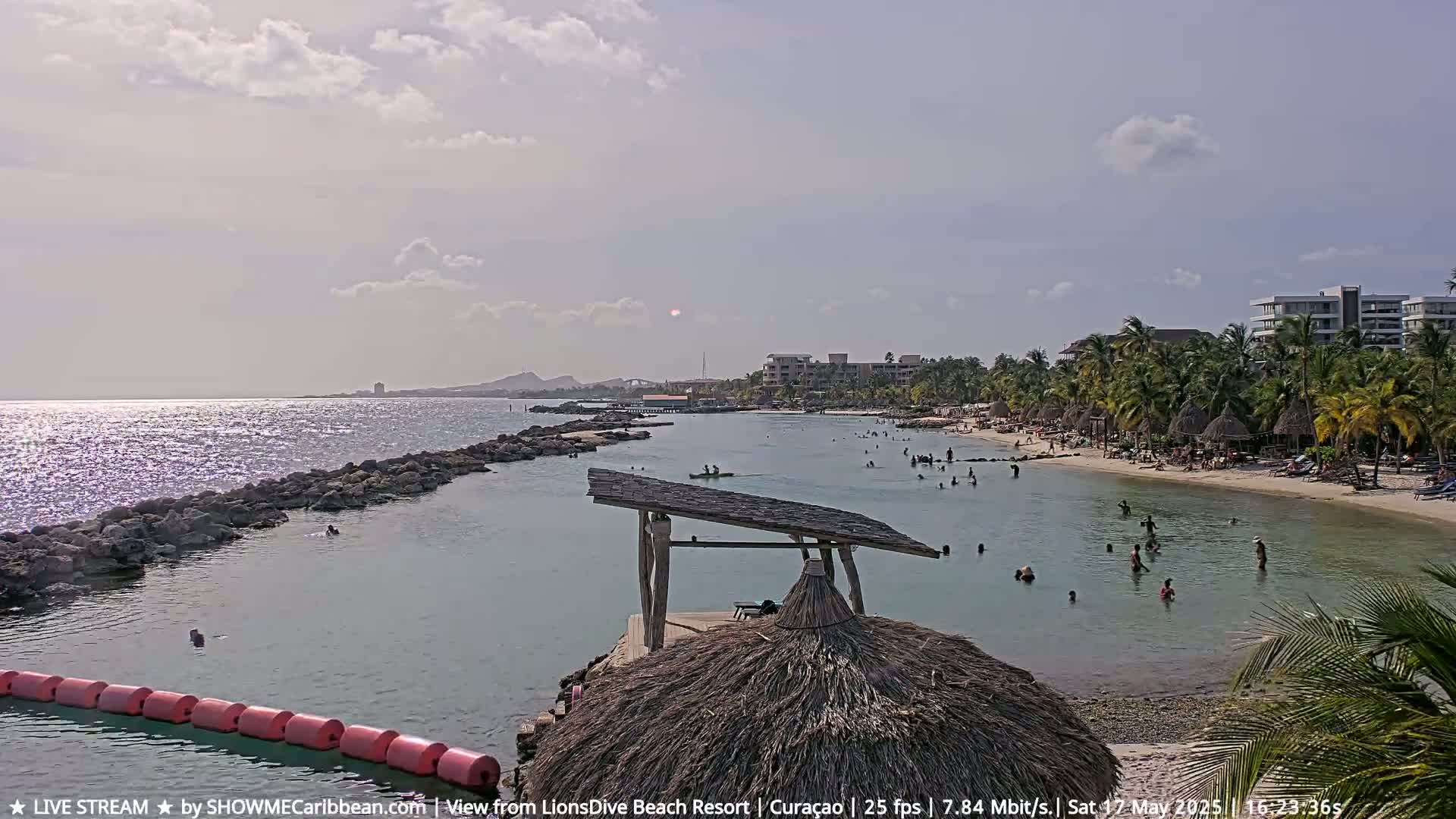 A partly sunny day at a calm, shallow beach with many people swimming and lounging, featuring a thatched-roof hut and a rock jetty.