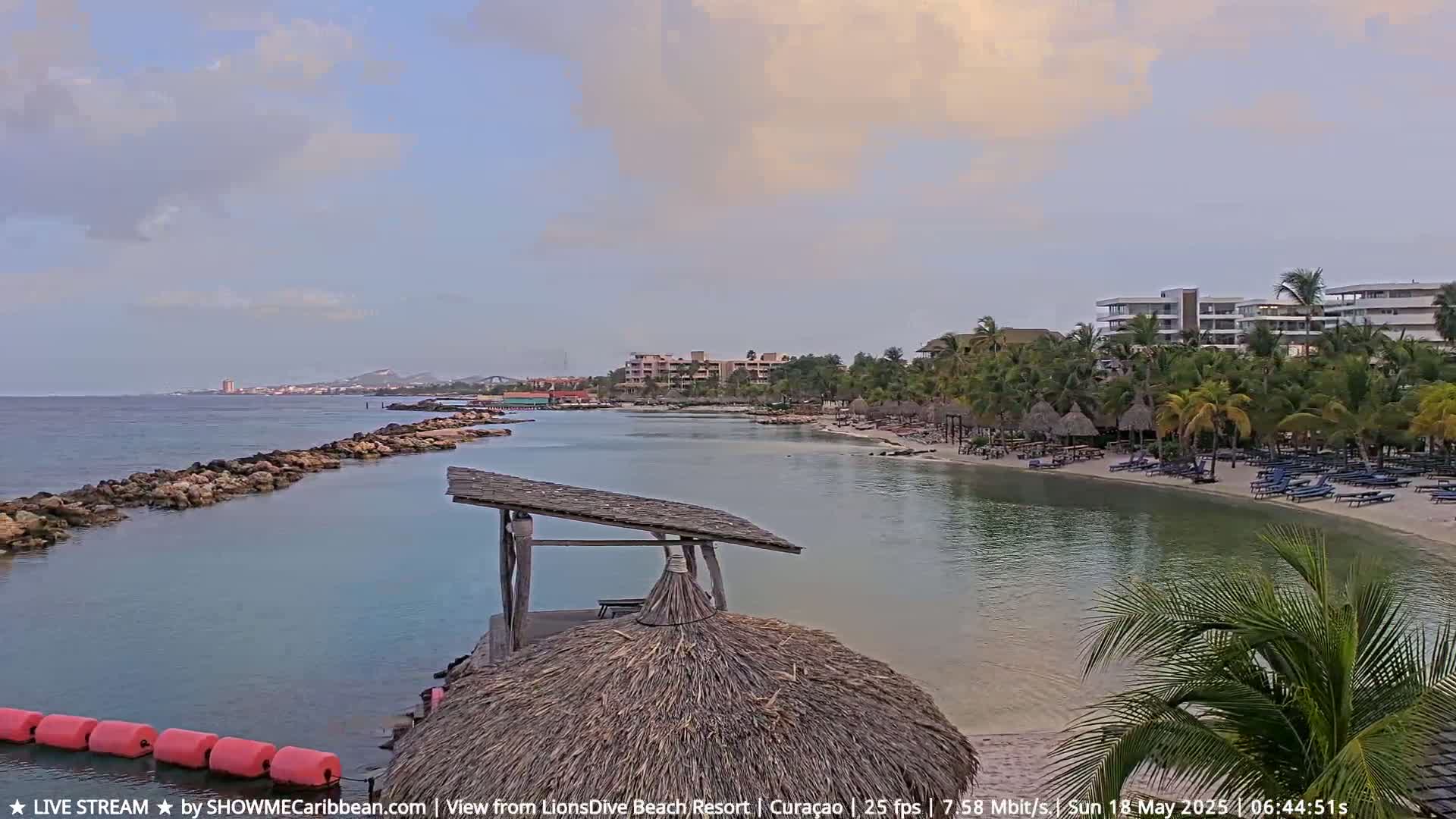 A partly cloudy day reveals a tranquil beach scene with calm, shallow turquoise water, a sandy shore lined with thatched huts and lounge chairs, and resort buildings in the background.