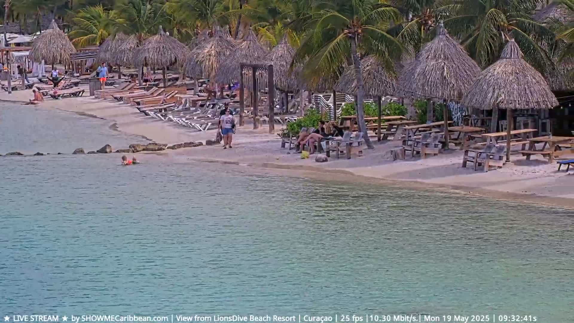 A sandy beach with numerous thatched umbrellas, lounge chairs, and picnic tables is partially surrounded by calm, clear turquoise water, and people are relaxing and walking around the area.