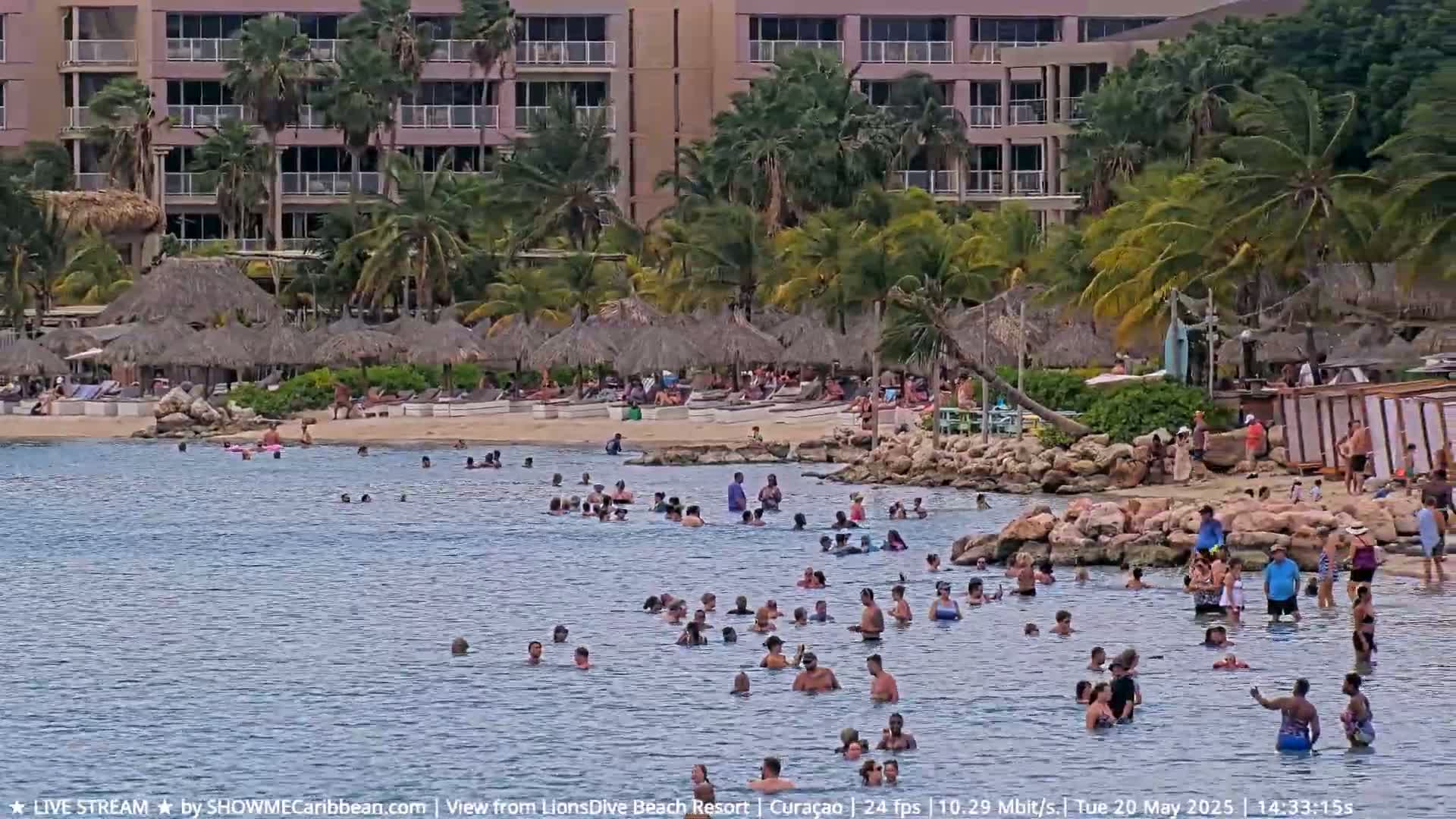 Many people are swimming and relaxing on a beach near a resort with palm trees under a partly cloudy sky.
