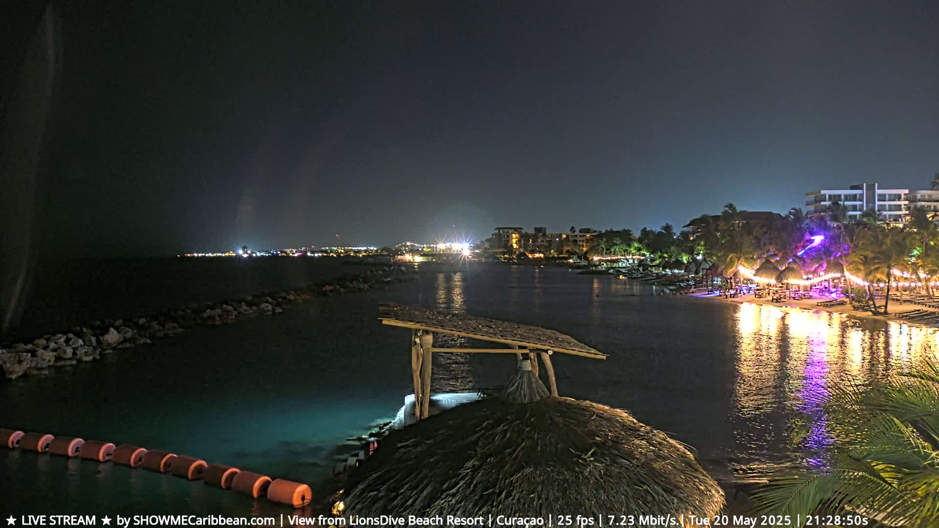 A nighttime view from a thatched roof structure shows a calm body of water reflecting lights from a beachside resort and buildings across the water under a dark sky.