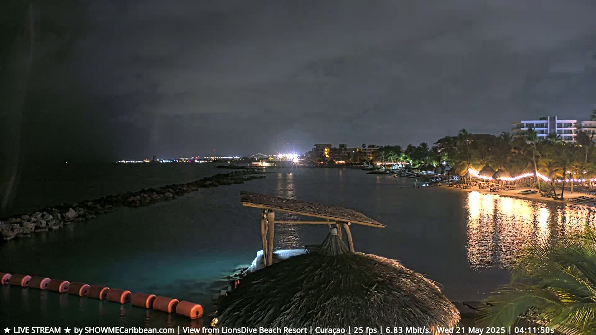 A nighttime view of a calm bay, illuminated by lights from a nearby resort and string lights on the shore under a dark, cloudy sky.