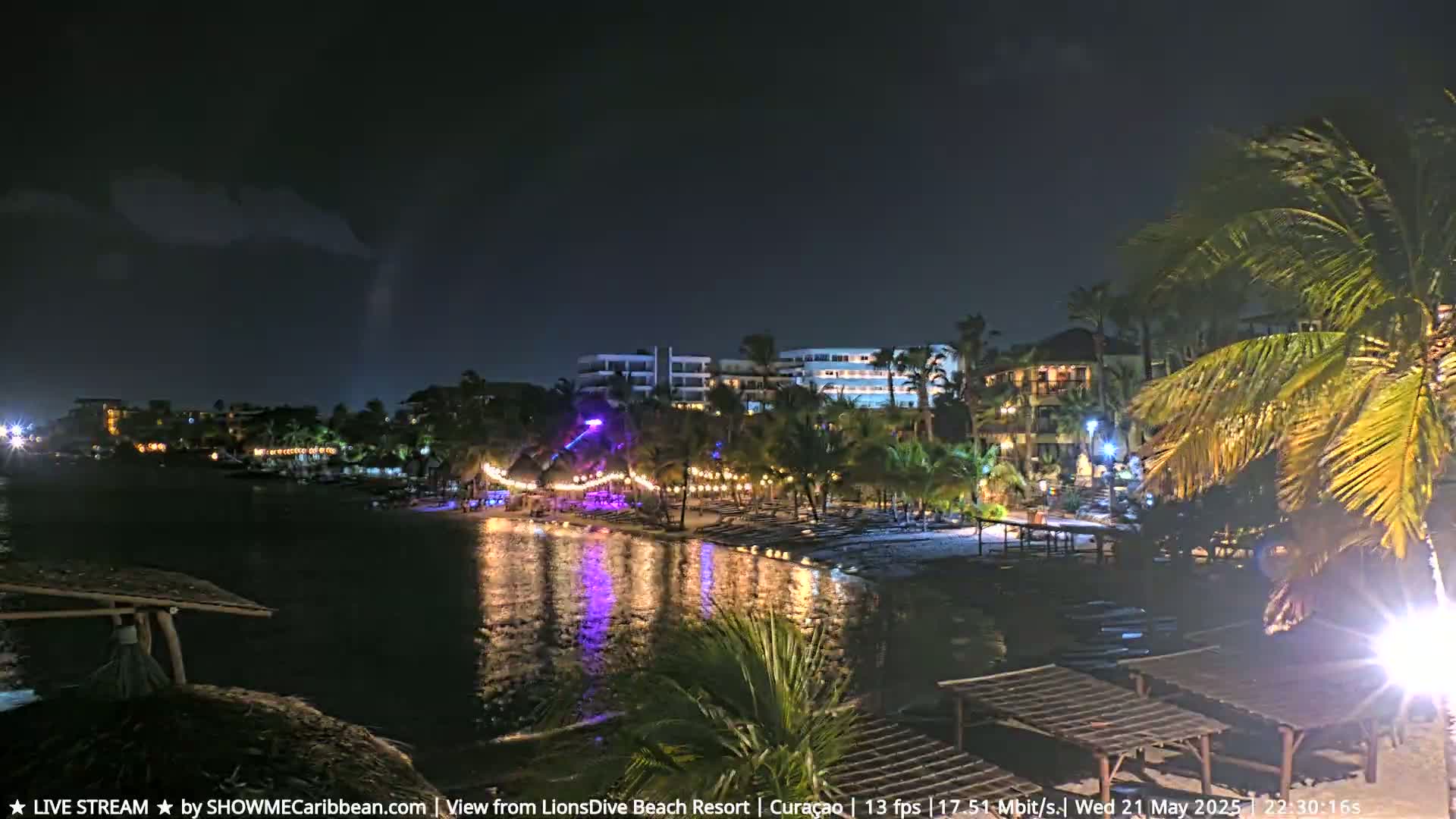 A nighttime view of a resort beach with calm water reflecting lights from buildings and string lights along the shore under a mostly clear sky.