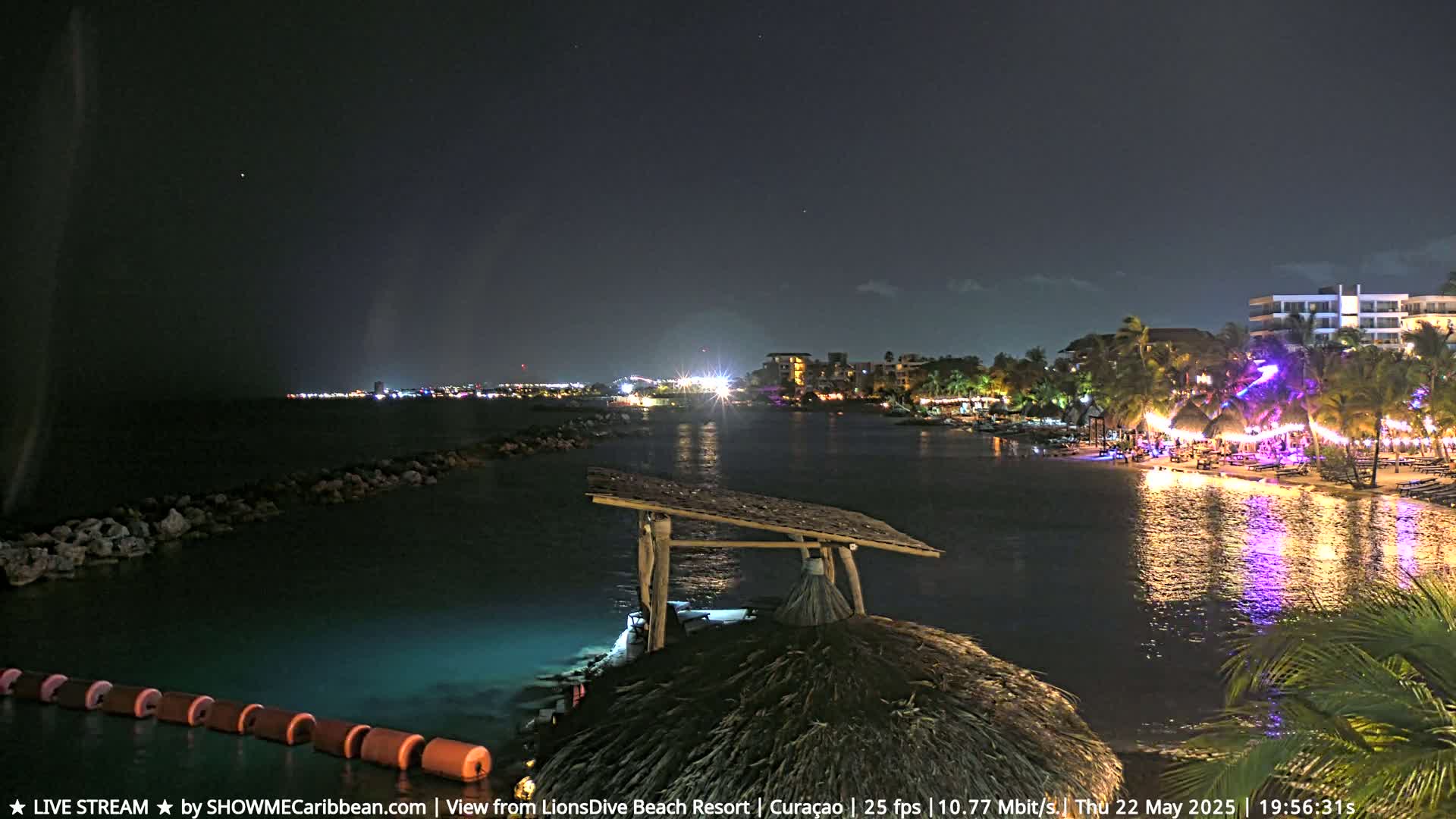 A nighttime view of a calm bay with a brightly lit beachfront resort and a thatched roof structure in the foreground under a clear, starry sky.