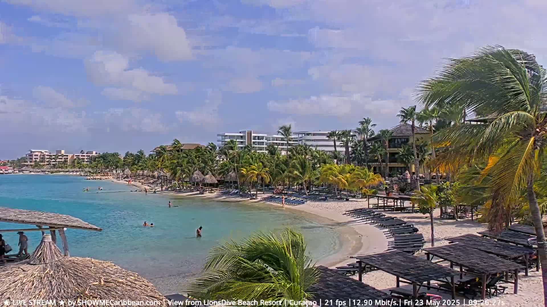 A partly sunny day on a tropical beach with calm turquoise water, palm trees, beach chairs, and resort buildings in the background.