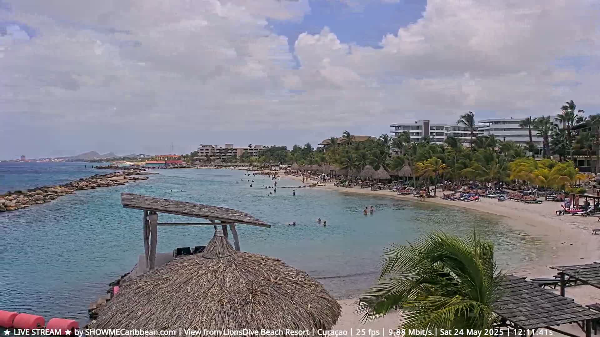 A partly cloudy day at a tropical beach with calm turquoise water, a sandy shore lined with palm trees, beach umbrellas, and buildings.