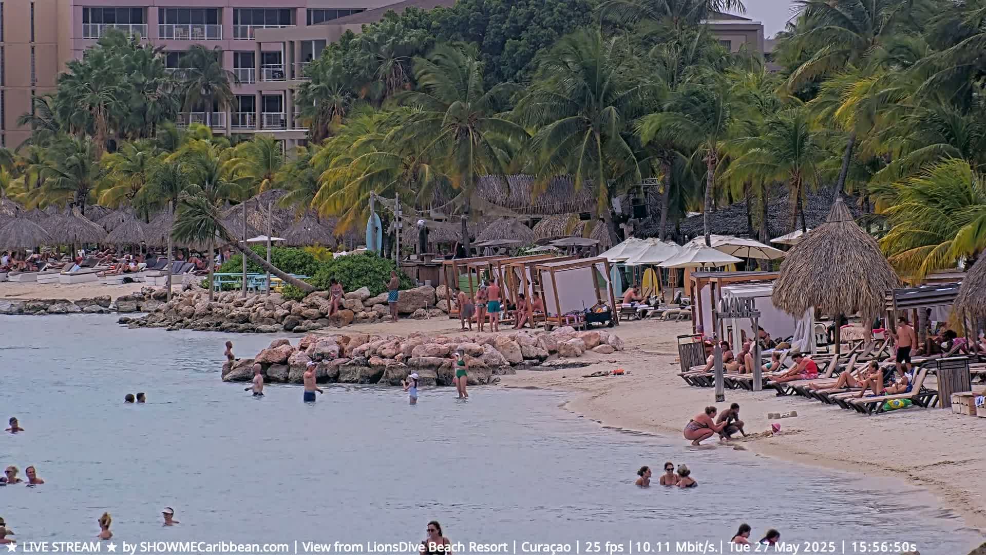 A sandy beach with many people swimming, sunbathing, and relaxing under umbrellas and shaded structures, next to a calm body of water, in the background are palm trees and buildings.