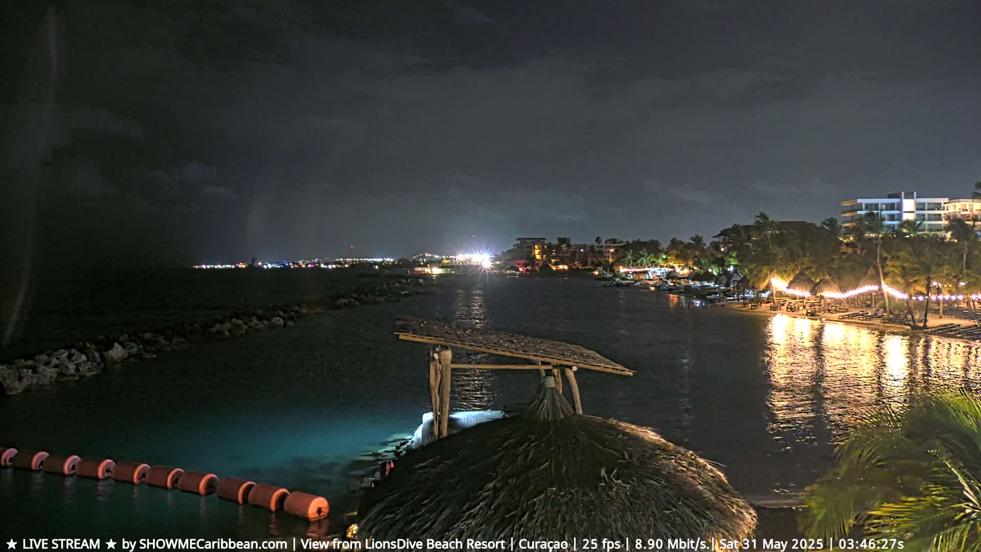 A nighttime view of a calm, illuminated body of water with a beach and buildings in the distance under a dark, cloudy sky.