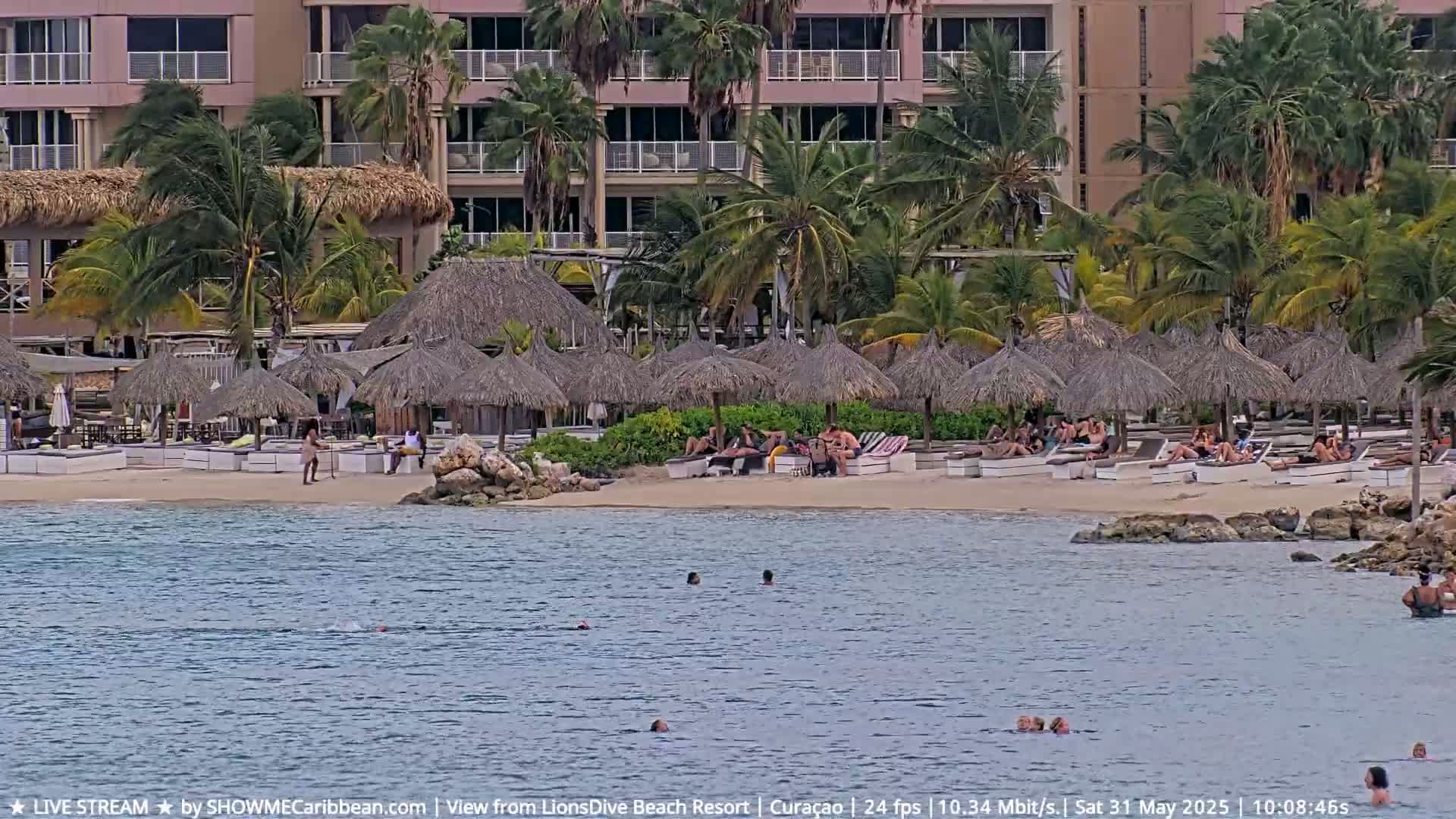 A sandy beach with thatched-roof umbrellas and people relaxing, swimming, and sunbathing, situated in front of a multi-story building, in breezy weather.