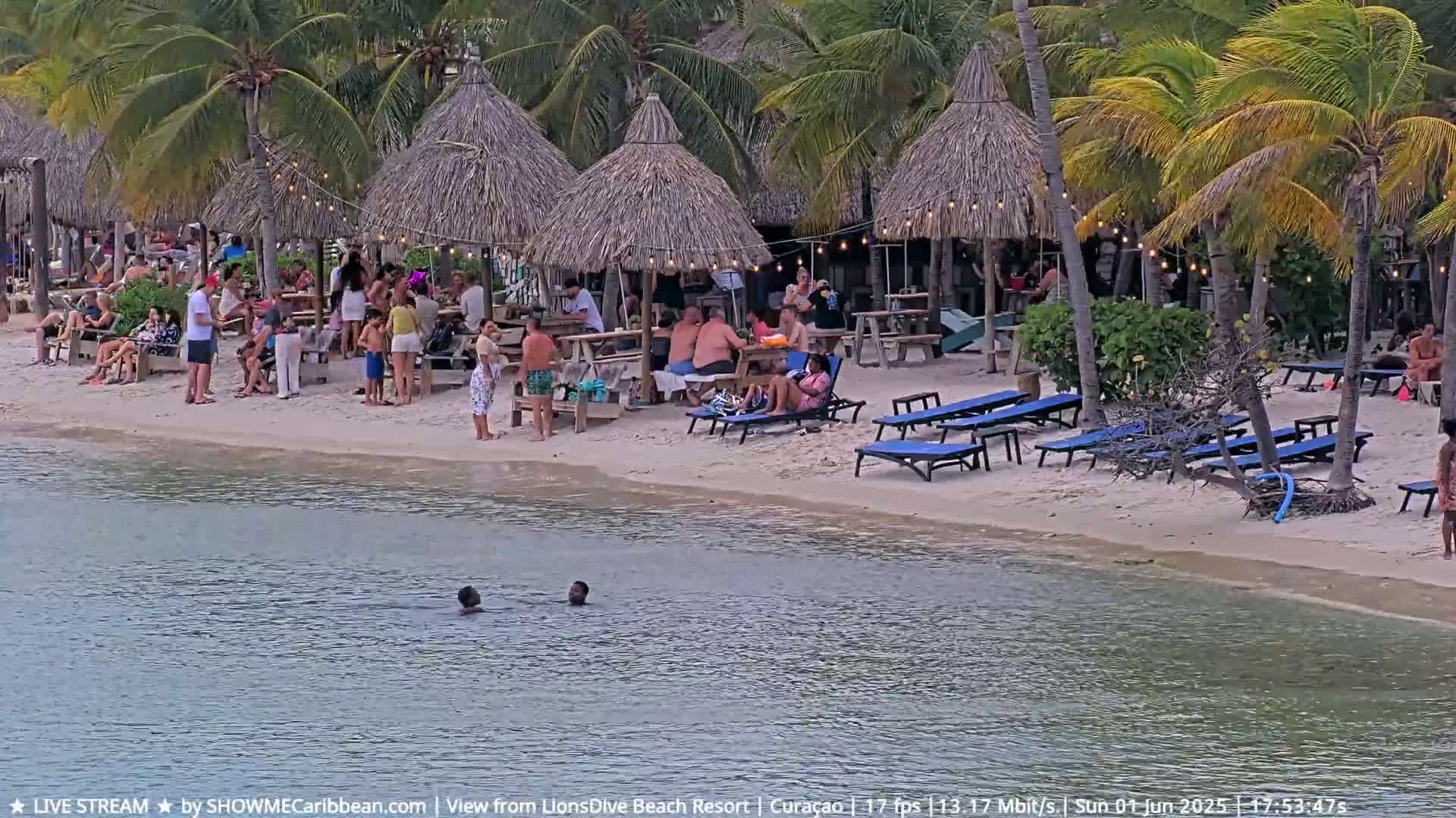 Many people relax on a sandy beach under thatched umbrellas near calm water, some swimming.
