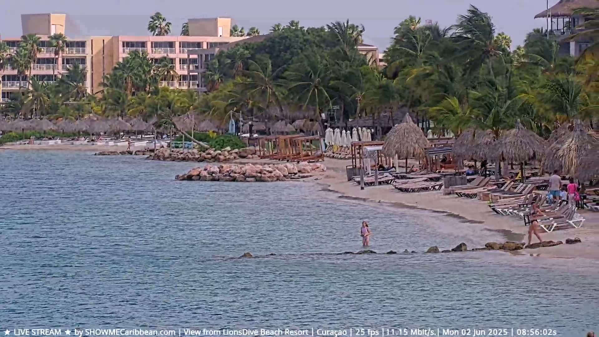 A sandy beach with thatched-roof umbrellas and lounge chairs, bordered by a calm ocean and a multi-story resort building behind it, under a partly cloudy sky with a slight wind.