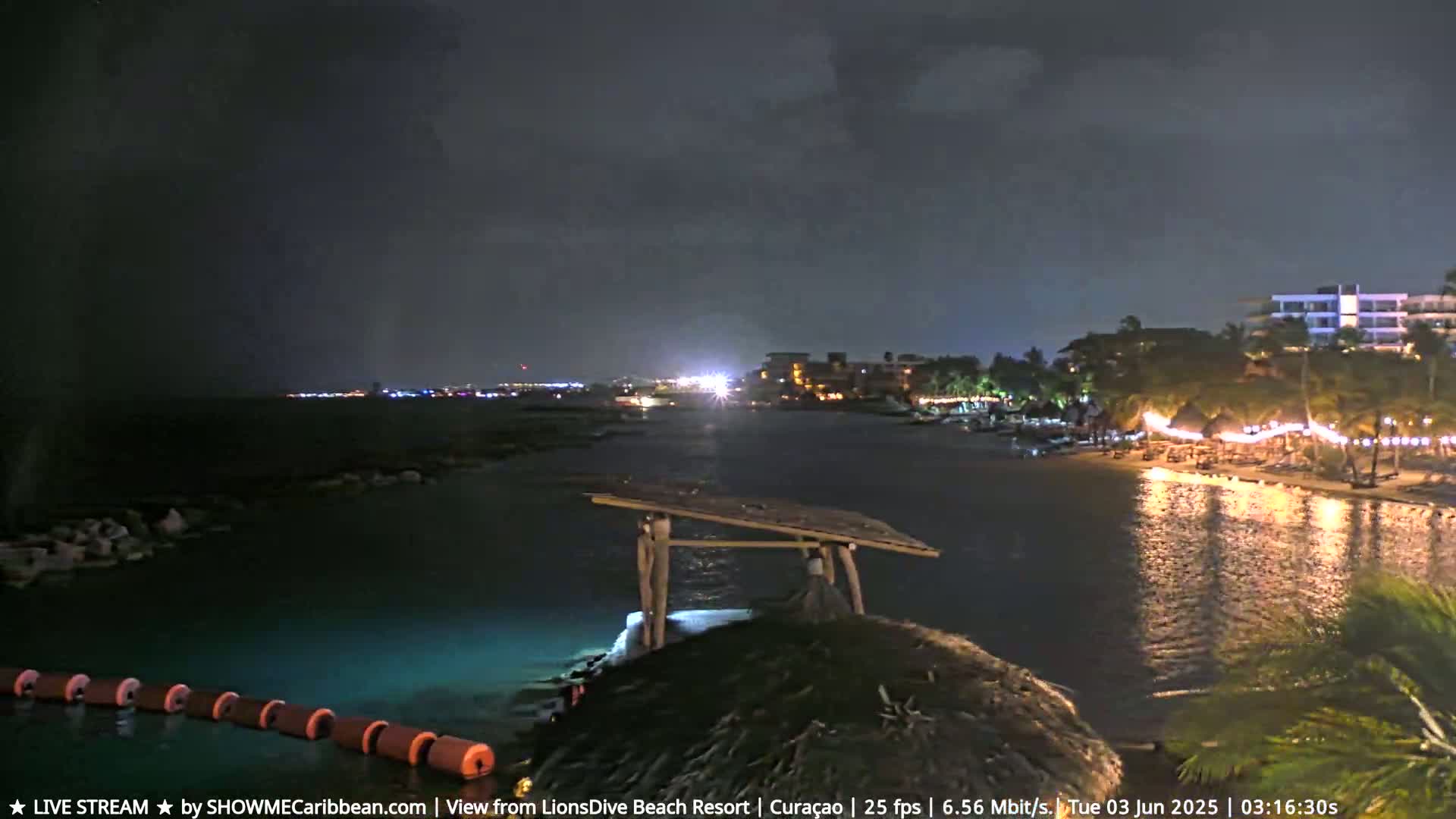 A nighttime coastal scene shows a calm body of water reflecting lights from a resort shore, with a simple wooden structure on a rocky outcrop in the foreground under a dark sky.