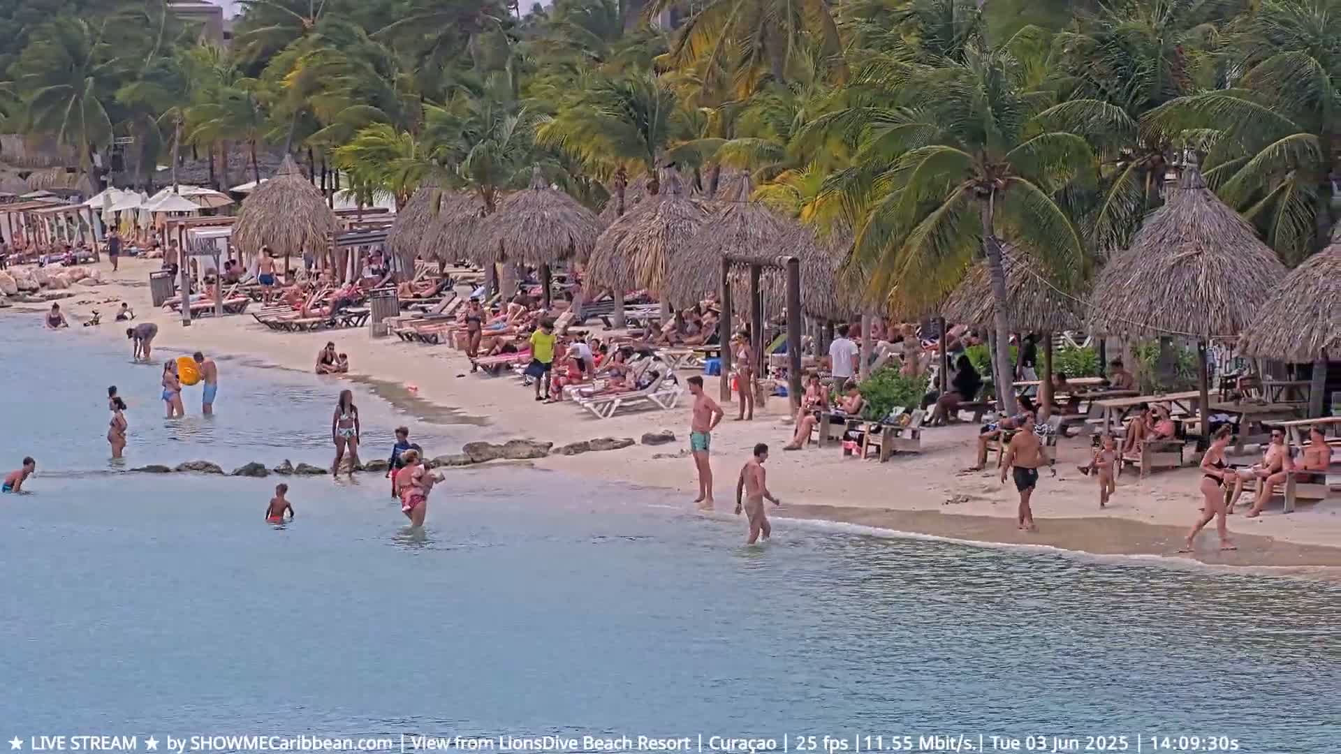 A sandy beach with many people swimming, sunbathing, and relaxing under thatched-roof umbrellas on a sunny day.