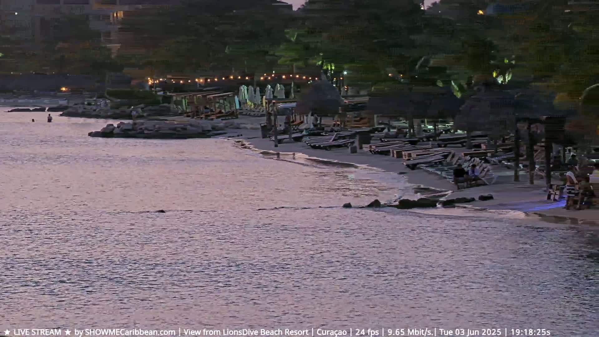 A beach at dusk shows calm water lapping at the shore near beach chairs and umbrellas under thatched roofs.