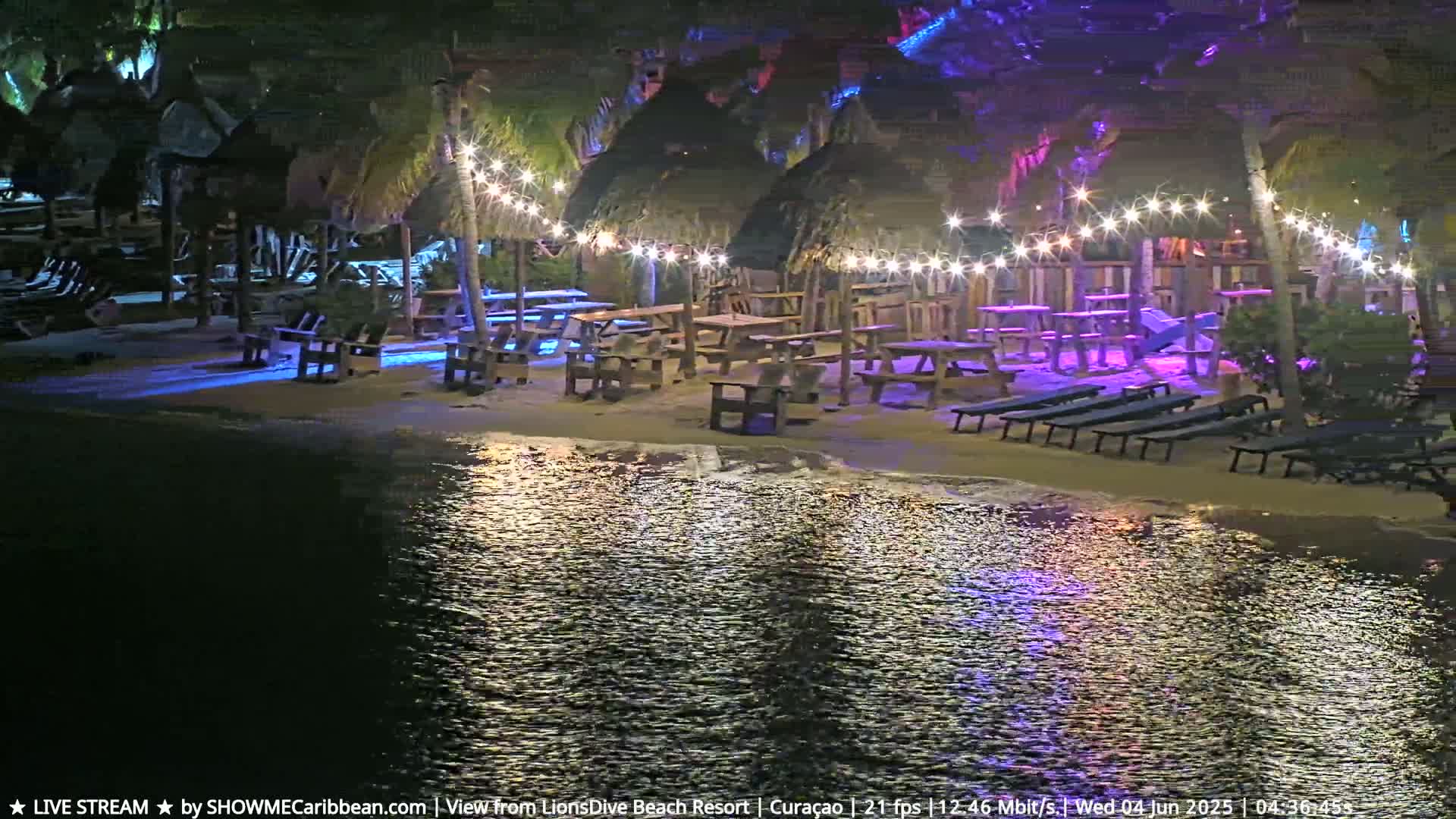 A nighttime beach scene shows picnic tables and lounge chairs under thatched-roof structures illuminated by string lights, with calm water reflecting the lights.