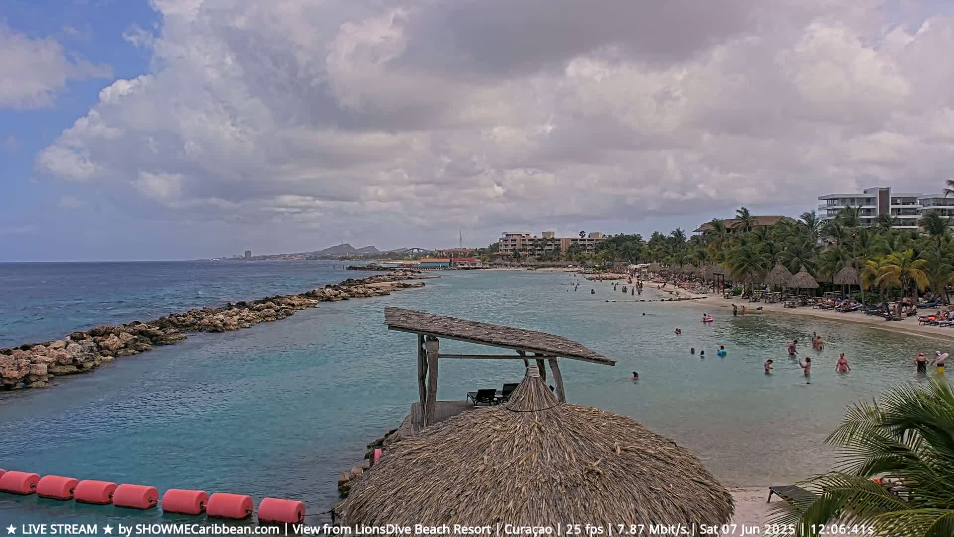 A partly cloudy day shows a calm, turquoise ocean with a rocky shoreline leading to a sandy beach where many people are swimming and relaxing under palm trees near resort buildings.
