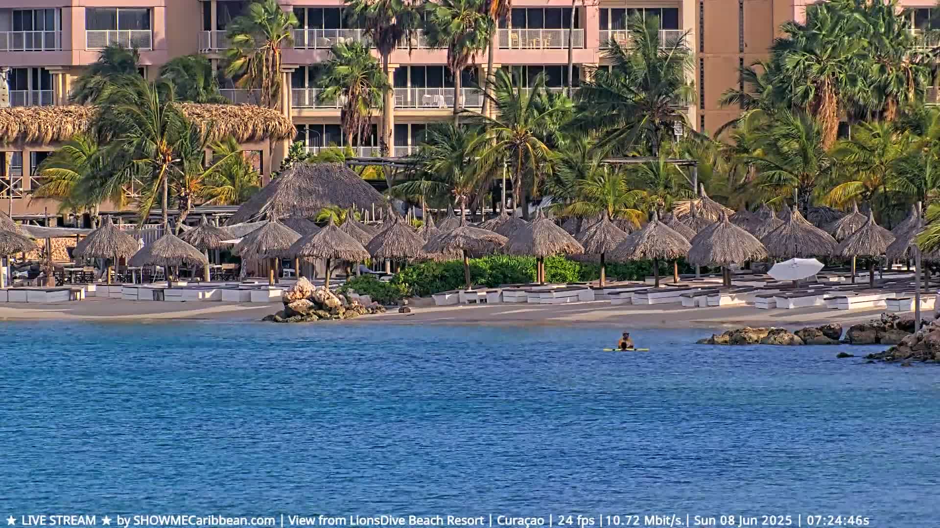 A sandy beach with numerous thatched umbrellas and lounge chairs is situated in front of a multi-story building, all surrounded by lush tropical vegetation, and a person is seen in the calm, blue ocean water.