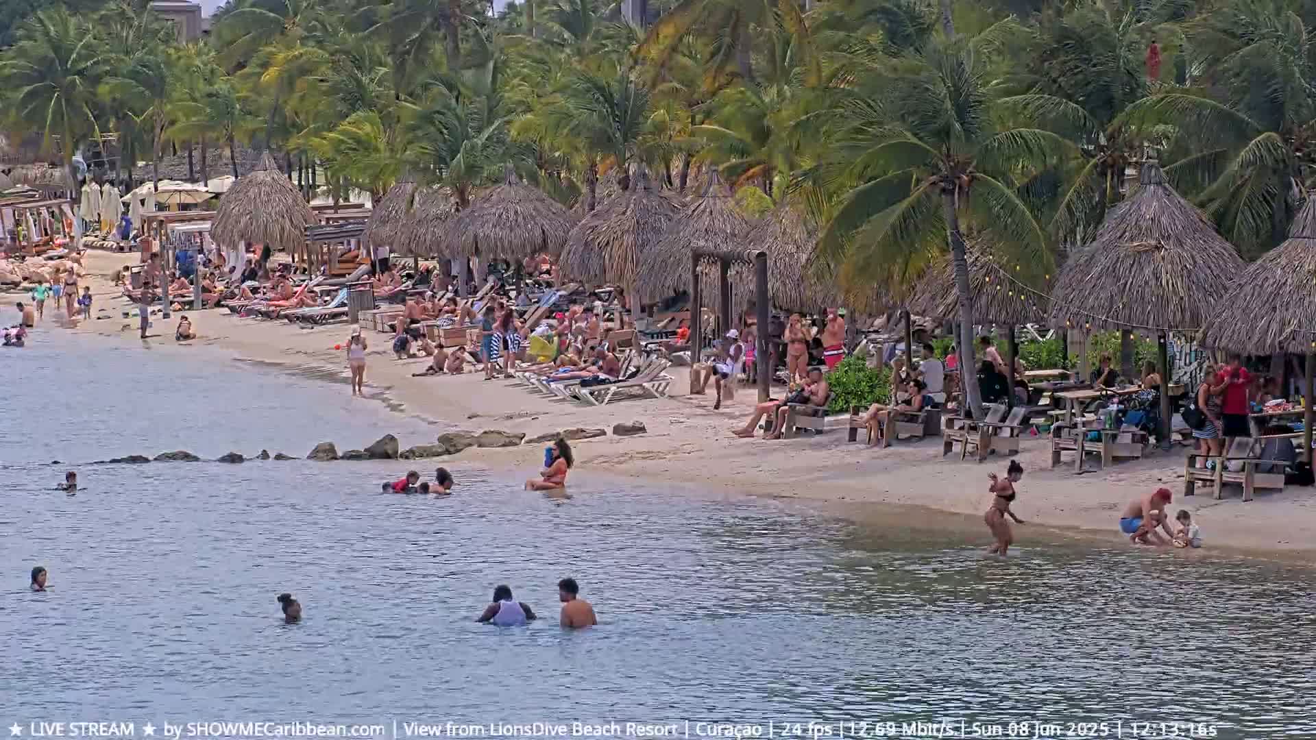 A sandy beach with numerous palm trees and thatched-roof structures is crowded with people swimming and lounging in calm water under a partly sunny sky.