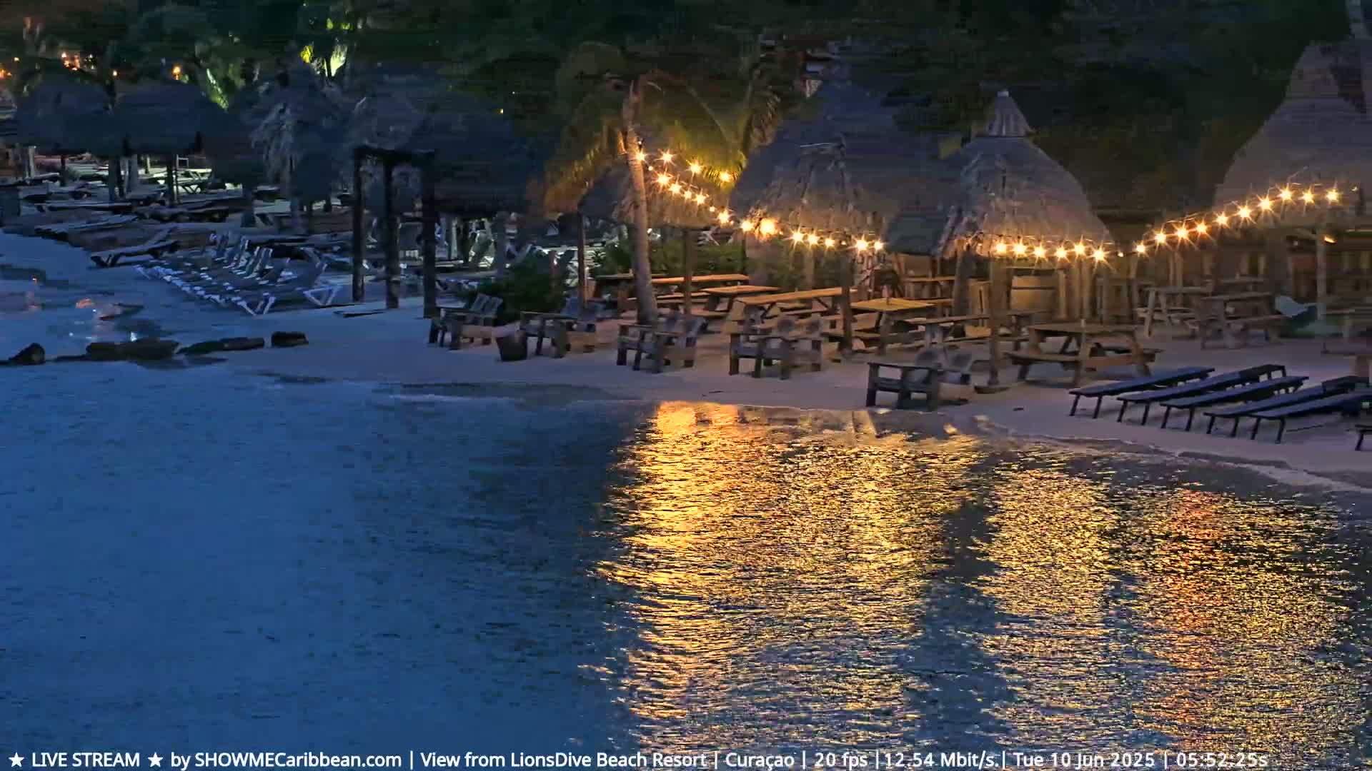 A beachside bar and restaurant with thatched-roof huts and string lights is illuminated at night, its lights reflecting in the calm water.