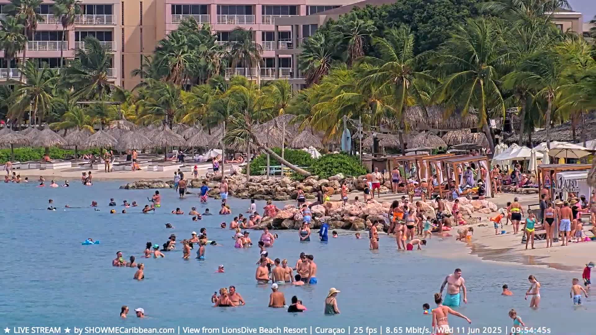 A crowded beach with many people swimming and relaxing in shallow water, under a sunny sky, near a resort with palm trees and thatched umbrellas.