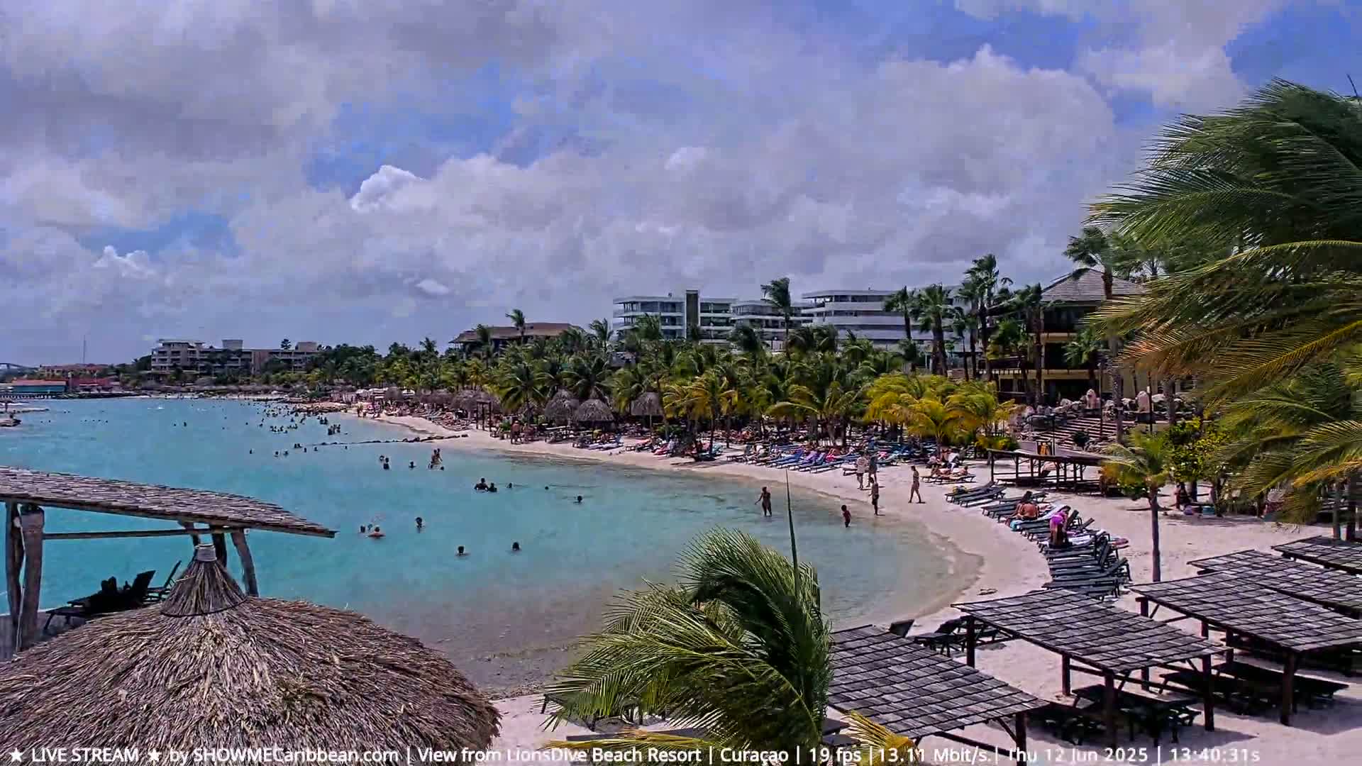 A partly cloudy day at a beach resort shows turquoise water with people swimming and sunbathing on the white sand beach, with palm trees and resort buildings in the background.