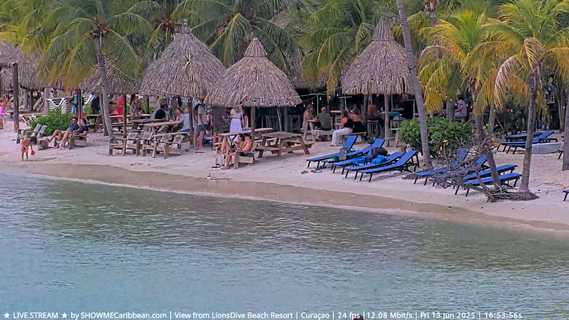 A sandy beach with thatch-roofed huts, picnic tables, lounge chairs, and people relaxing under a sunny sky next to calm water.