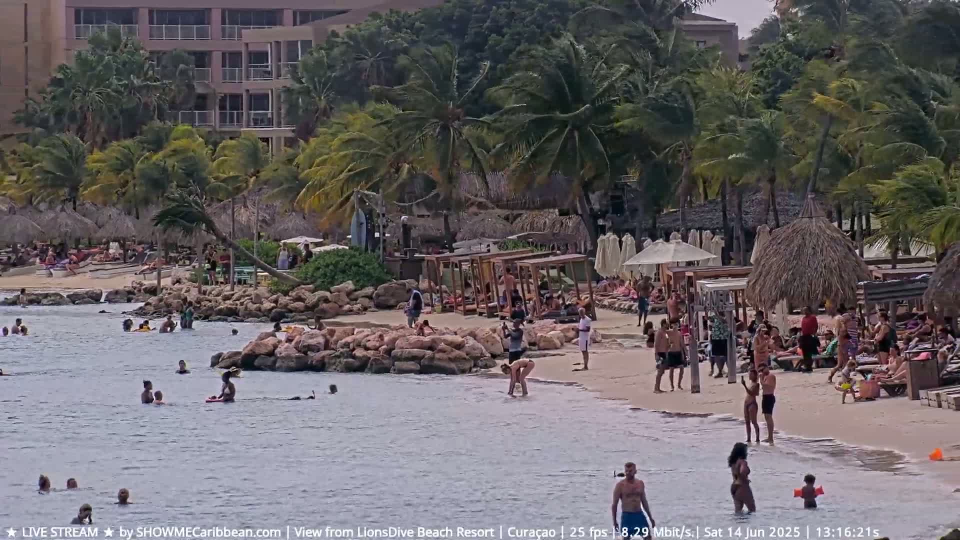 A sandy beach with many people swimming, sunbathing, and relaxing under beach umbrellas and shaded structures, all situated near a resort building under a partly cloudy sky.