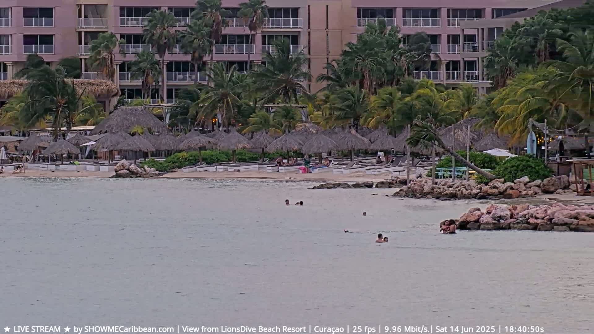 A sandy beach with many people swimming, sunbathing, and relaxing under beach umbrellas and shaded structures, all situated near a resort building under a partly cloudy sky.