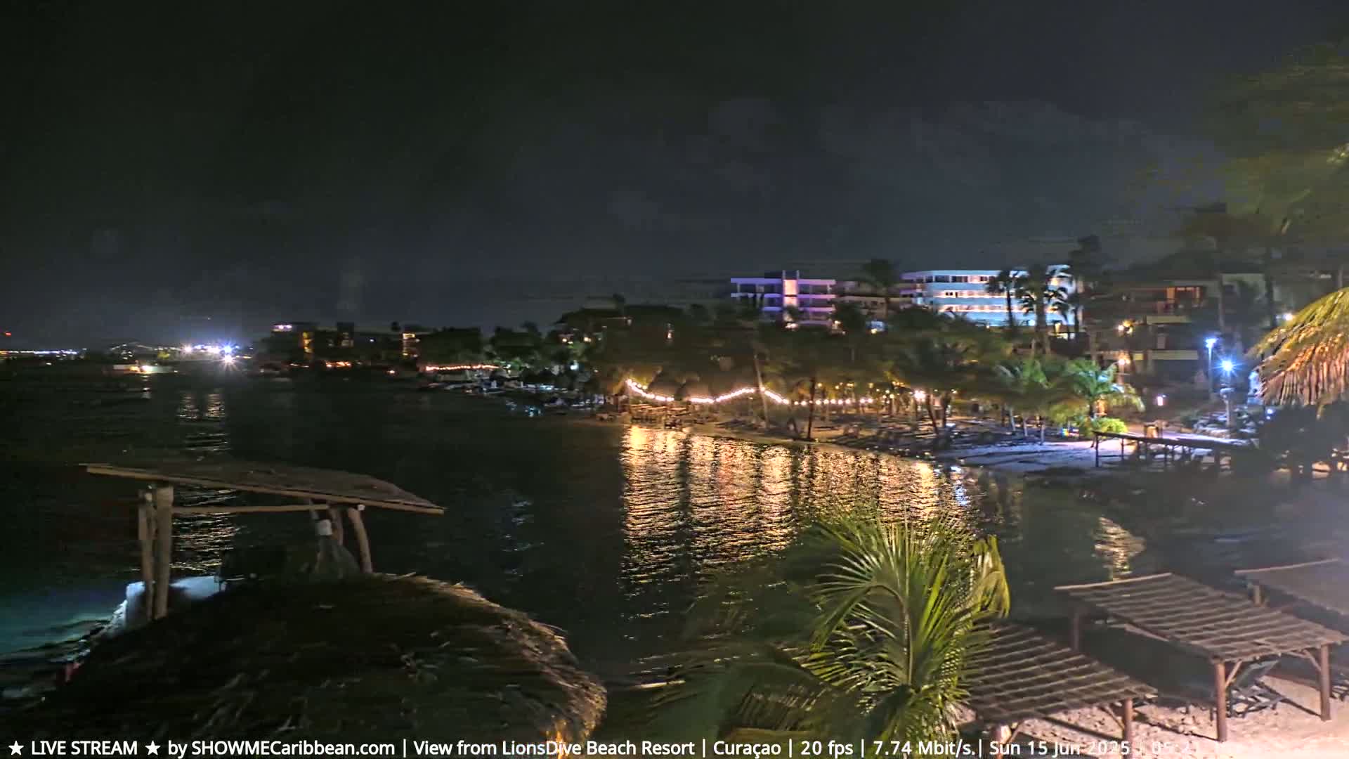 A nighttime view of a calm body of water reflecting lights from a beachside resort with palm trees and string lights.