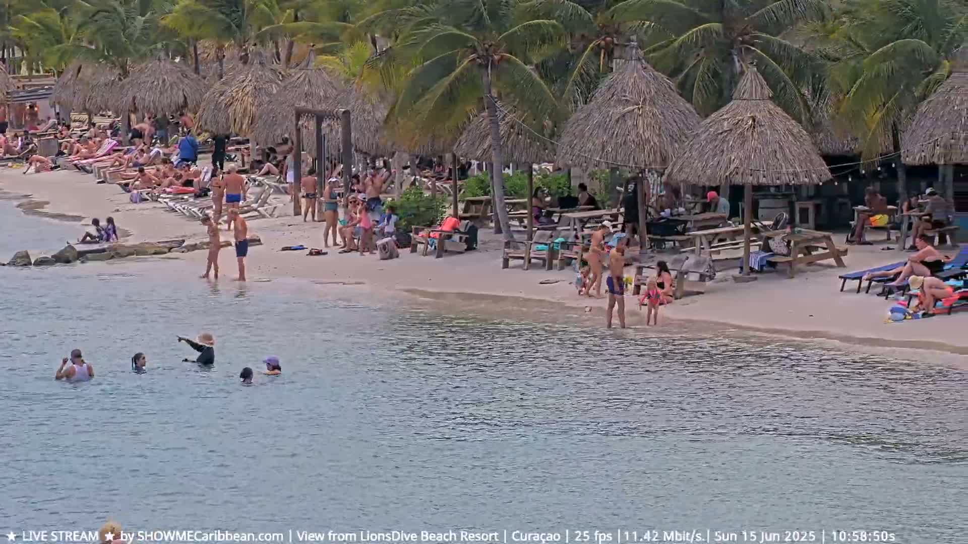 A sunny beach scene shows many people relaxing and swimming in calm water near thatched-roof beach huts.
