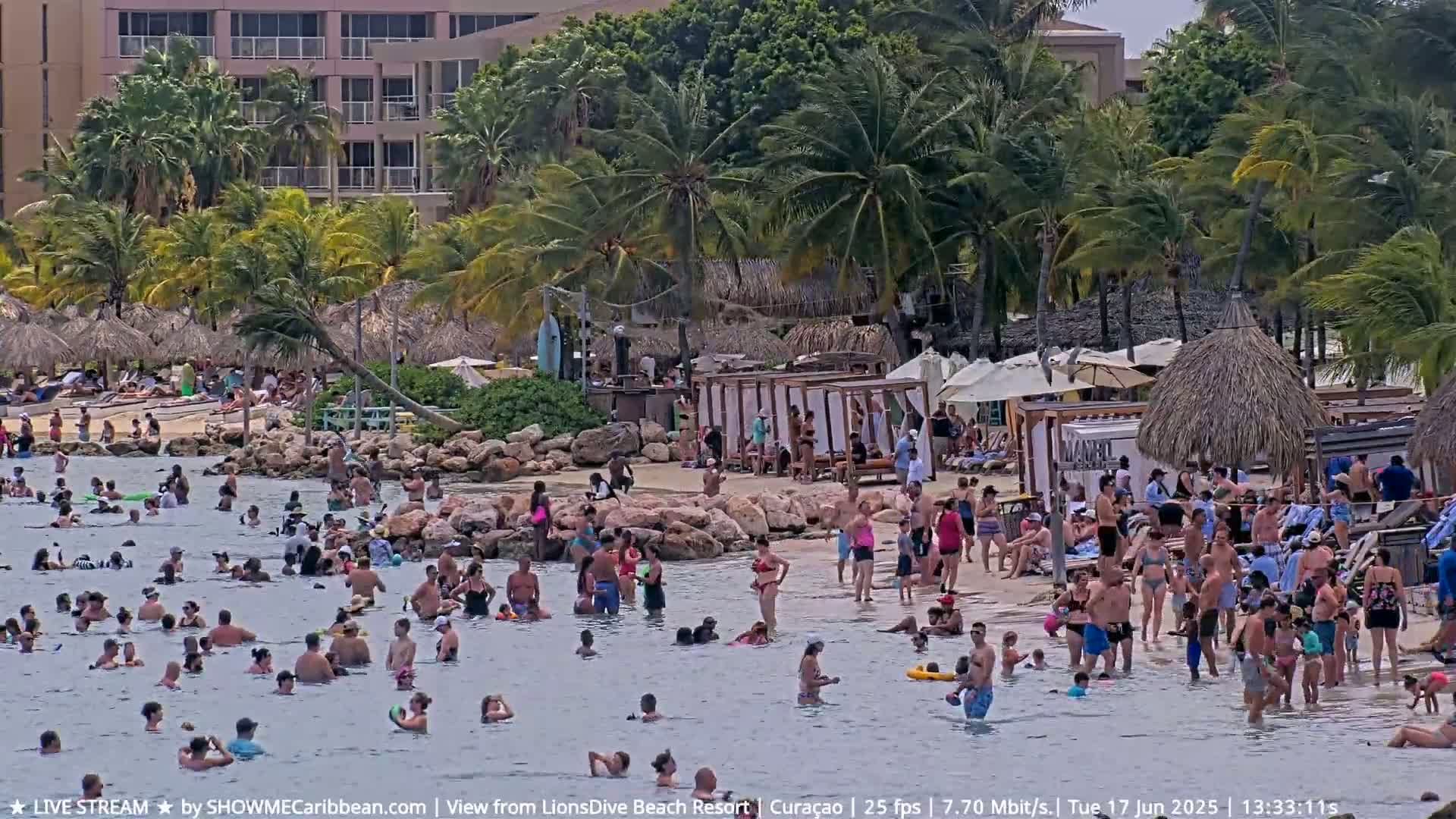 A crowded beach with many people swimming and sunbathing near a resort under a partly sunny sky.