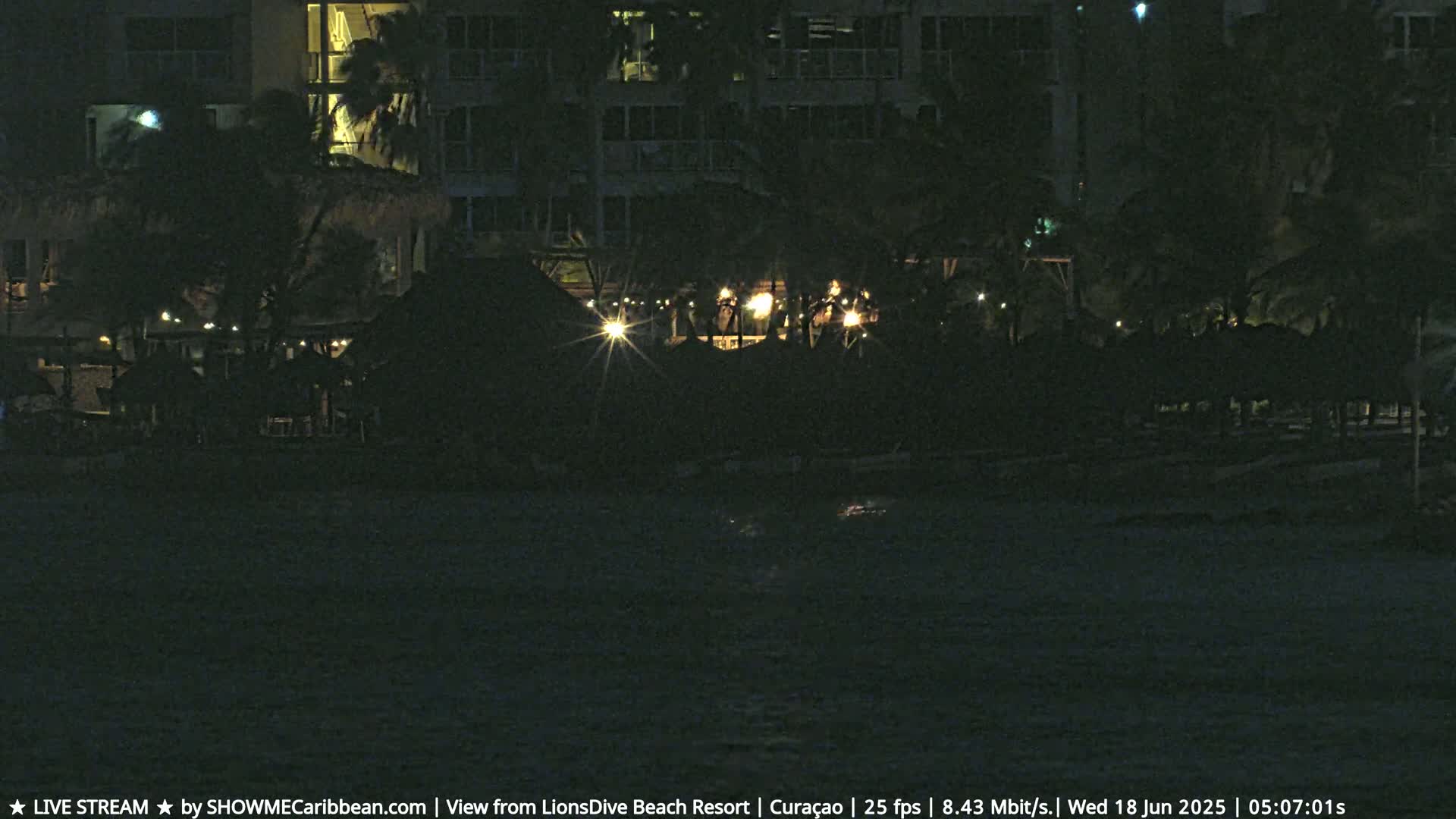 A dark nighttime scene shows a resort's beachfront area with dimly lit structures and palm trees near dark water.
