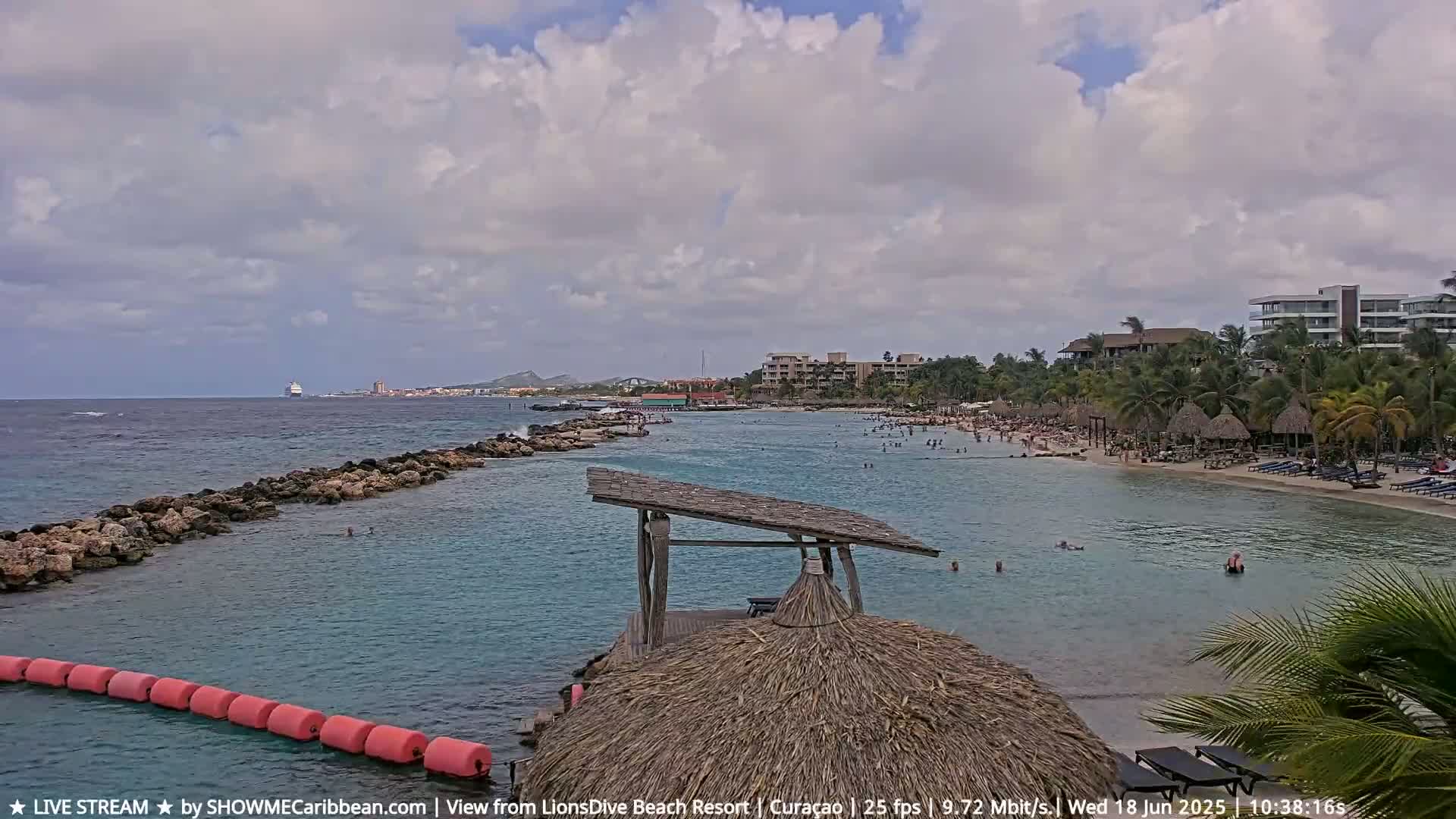 A partly cloudy day at a beach resort shows a calm, turquoise ocean with people swimming near a sandy shore lined with palm trees and buildings.