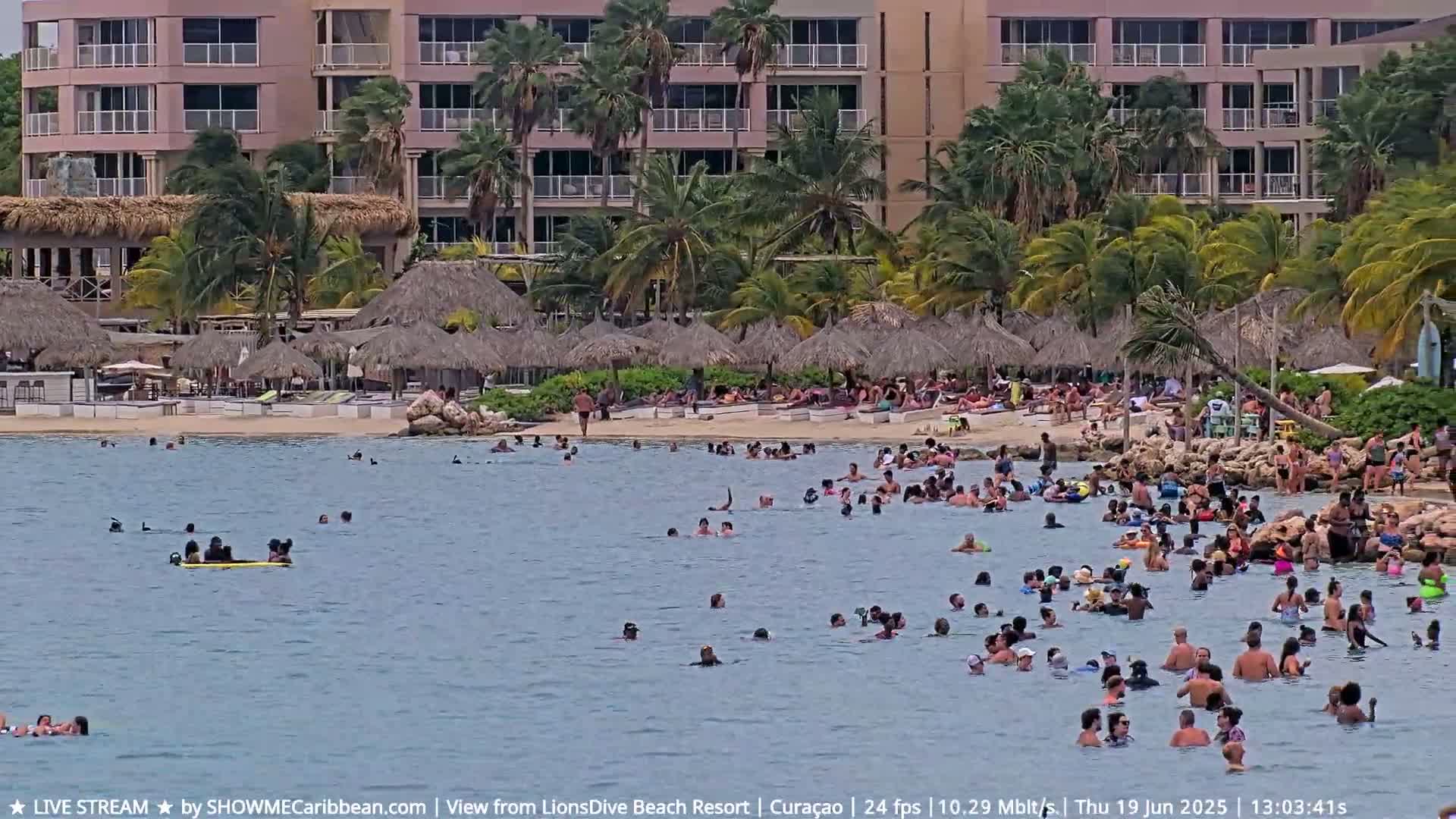A calm, sunny day at a beach resort shows many people swimming and relaxing in the water and on the sand, with thatch-roofed structures and a multi-story building in the background.