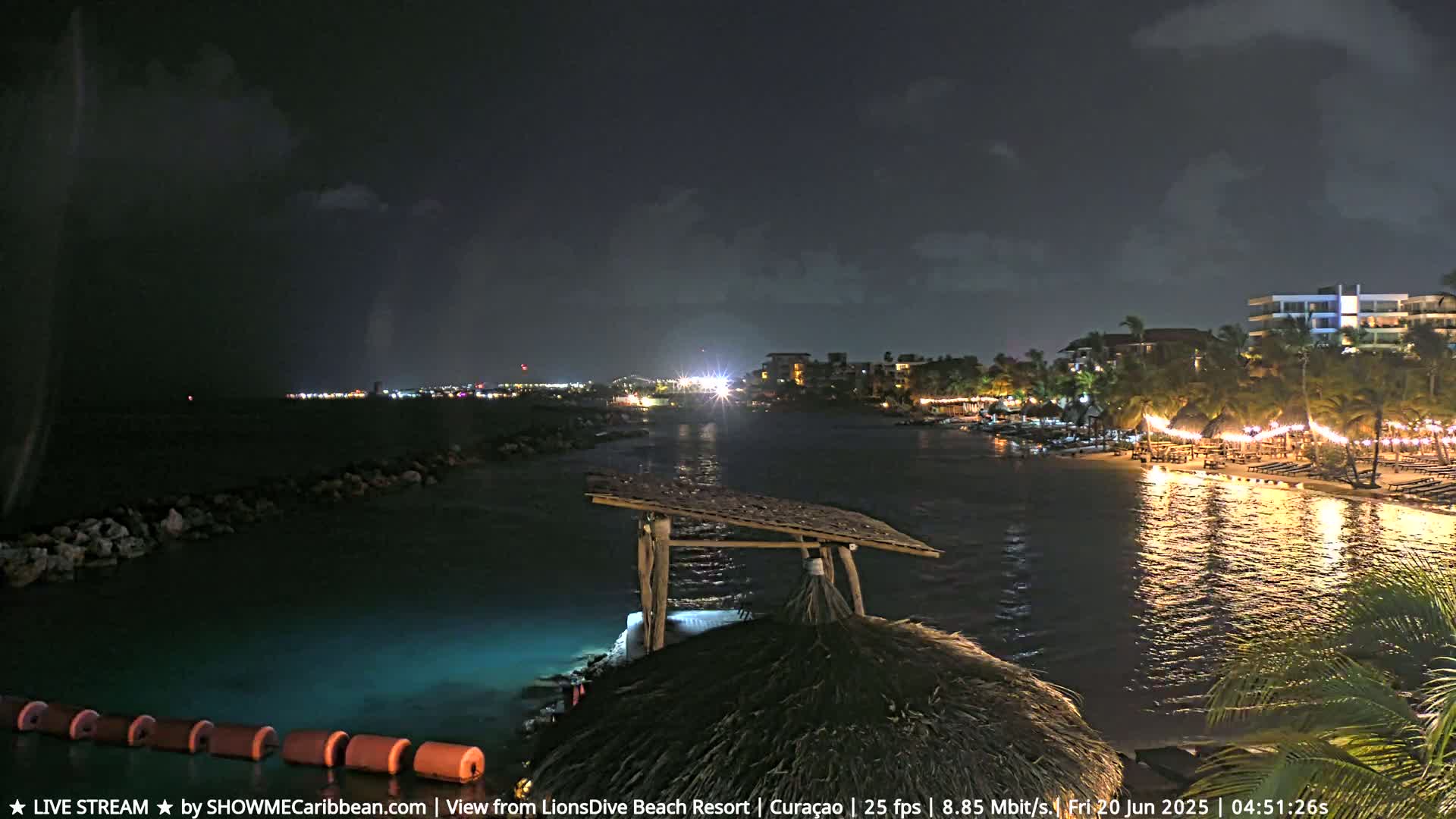 A nighttime view of a calm, lit-up beach and resort area along a body of water under a dark, mostly clear sky.