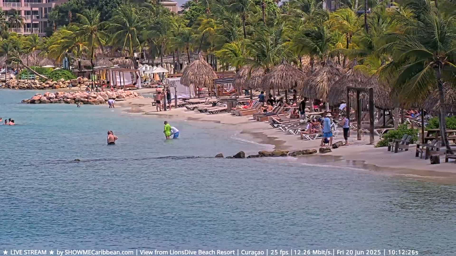 A sandy beach with palm trees, thatched-roof umbrellas, and lounge chairs is populated by people swimming and relaxing on a sunny day.