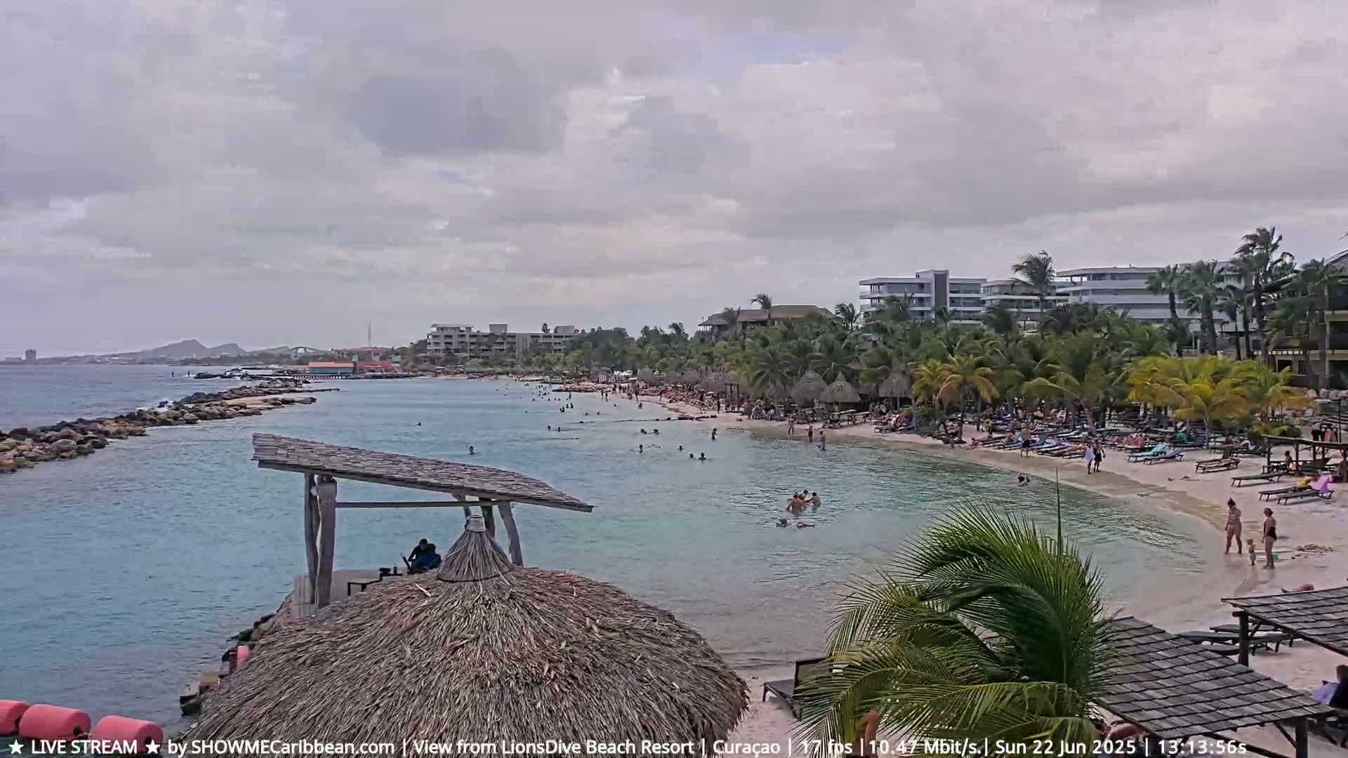 A sandy beach with numerous people swimming and relaxing under a partly cloudy sky.