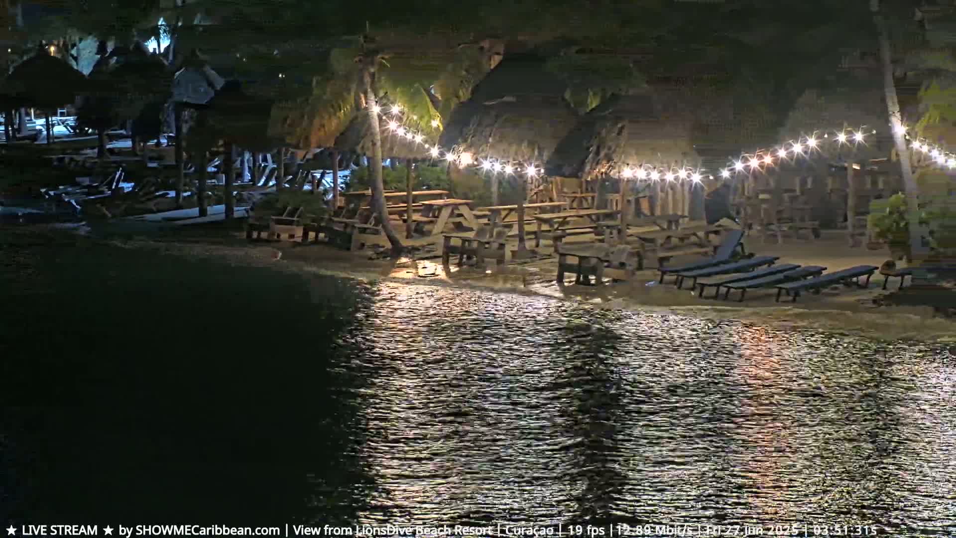 A nighttime scene shows a beachfront area with thatch-roofed structures, picnic tables, lounge chairs, and string lights illuminated against a calm body of water.